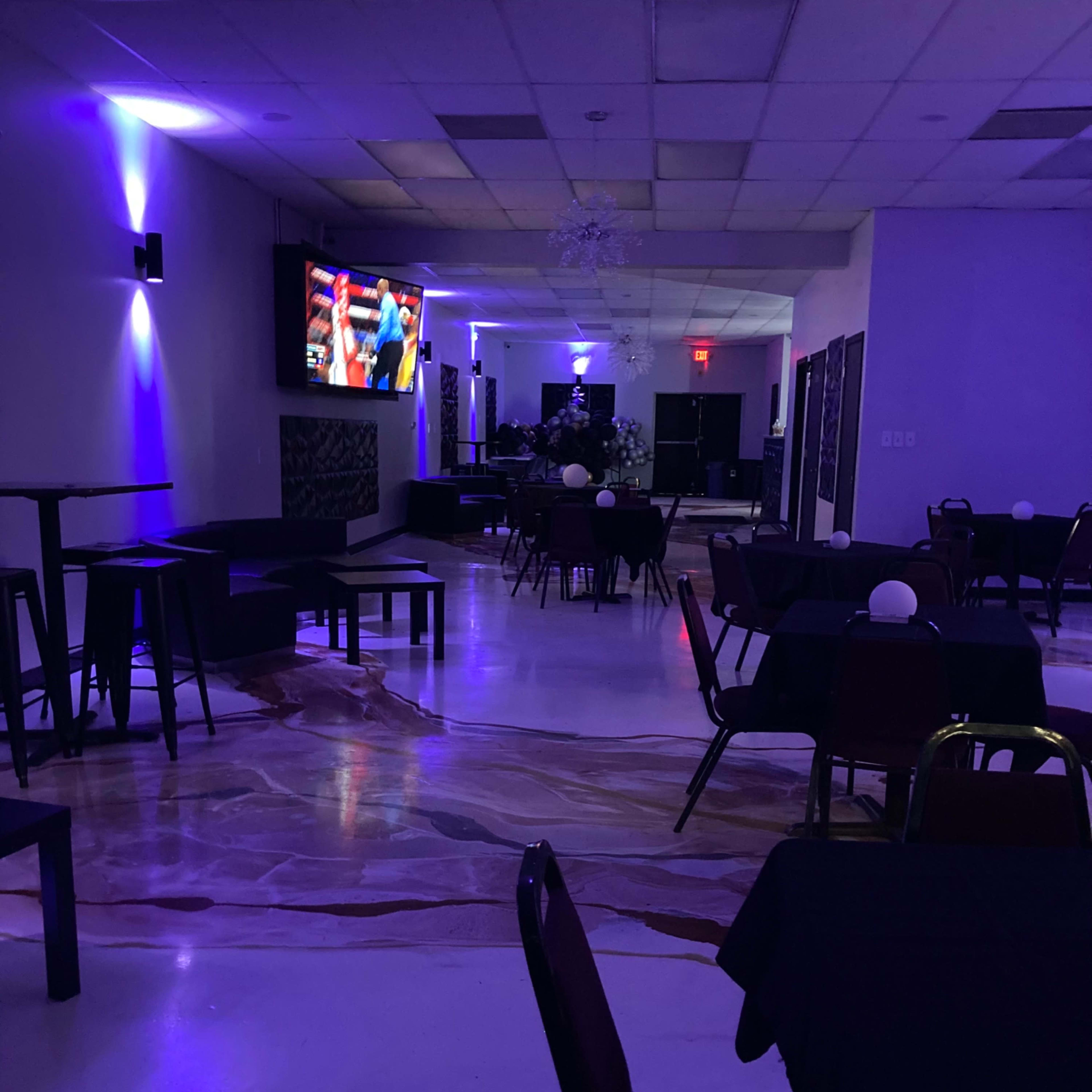 An empty restaurant with dark tables and chairs, illuminated by purple lighting and featuring a television screen displaying a sports event.