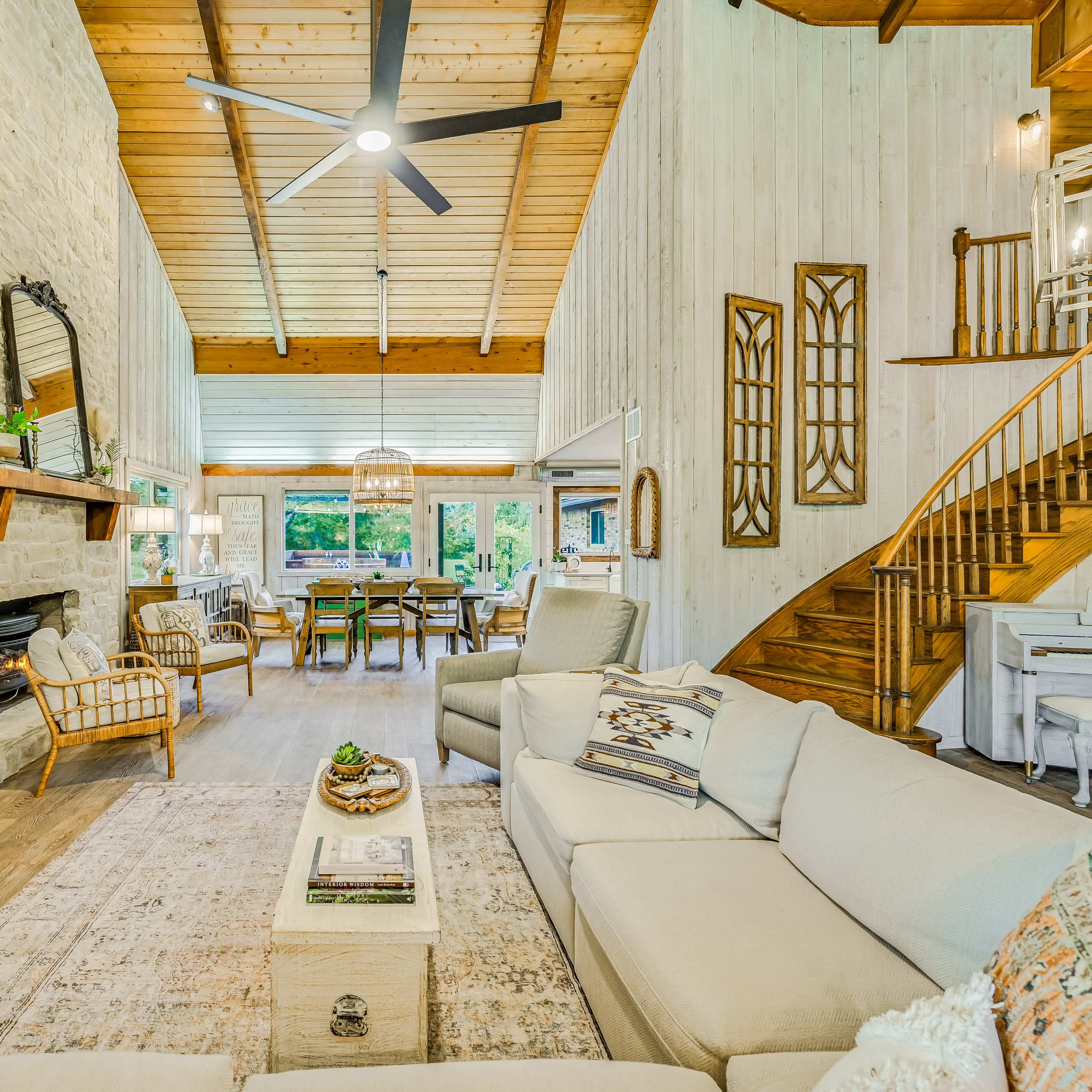 A spacious living room with high wooden ceilings, a stone fireplace, a large sofa, and a dining area visible through expansive windows.
