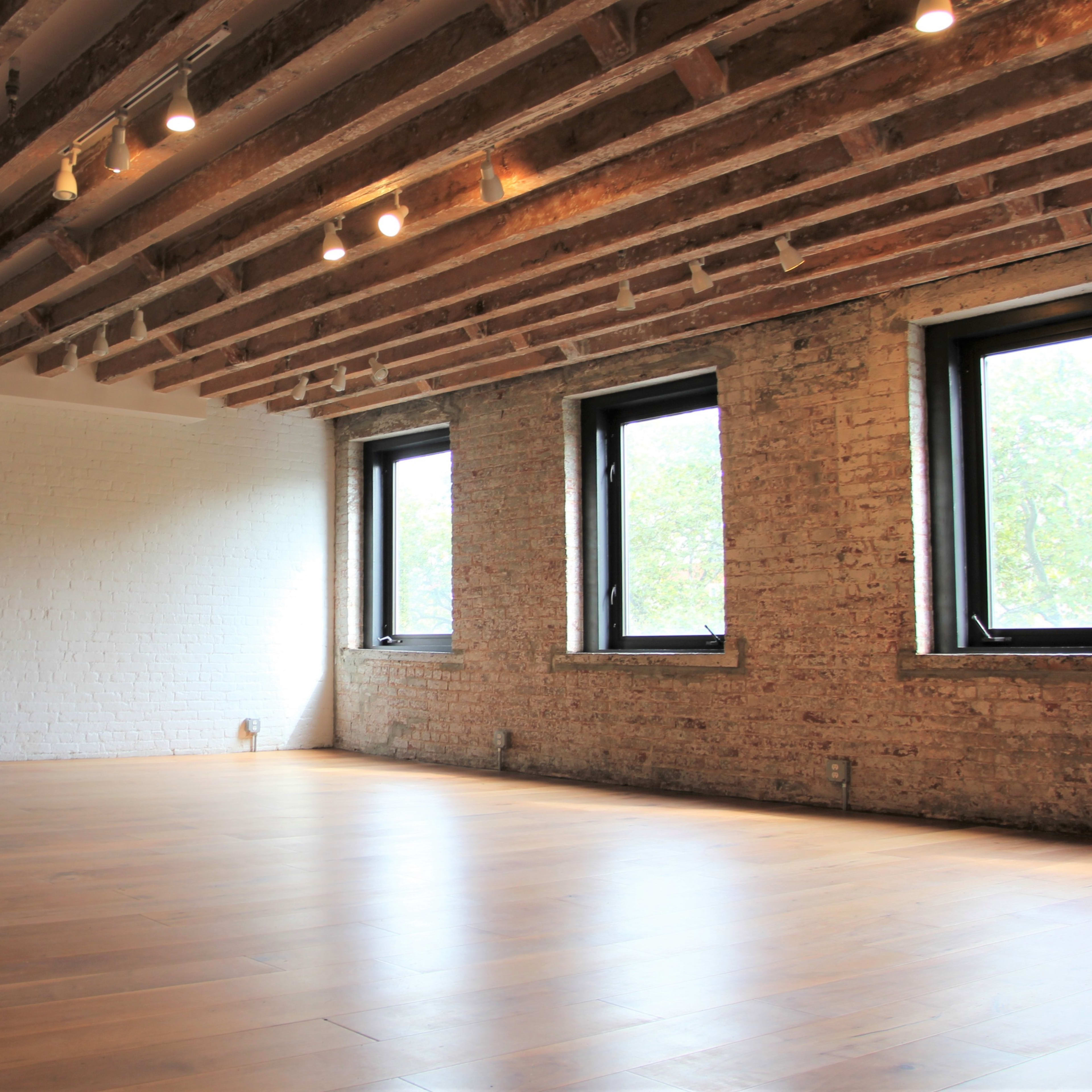 An empty room with wooden floorboards, exposed brick walls, and ceiling beams, featuring three windows that let in natural light.