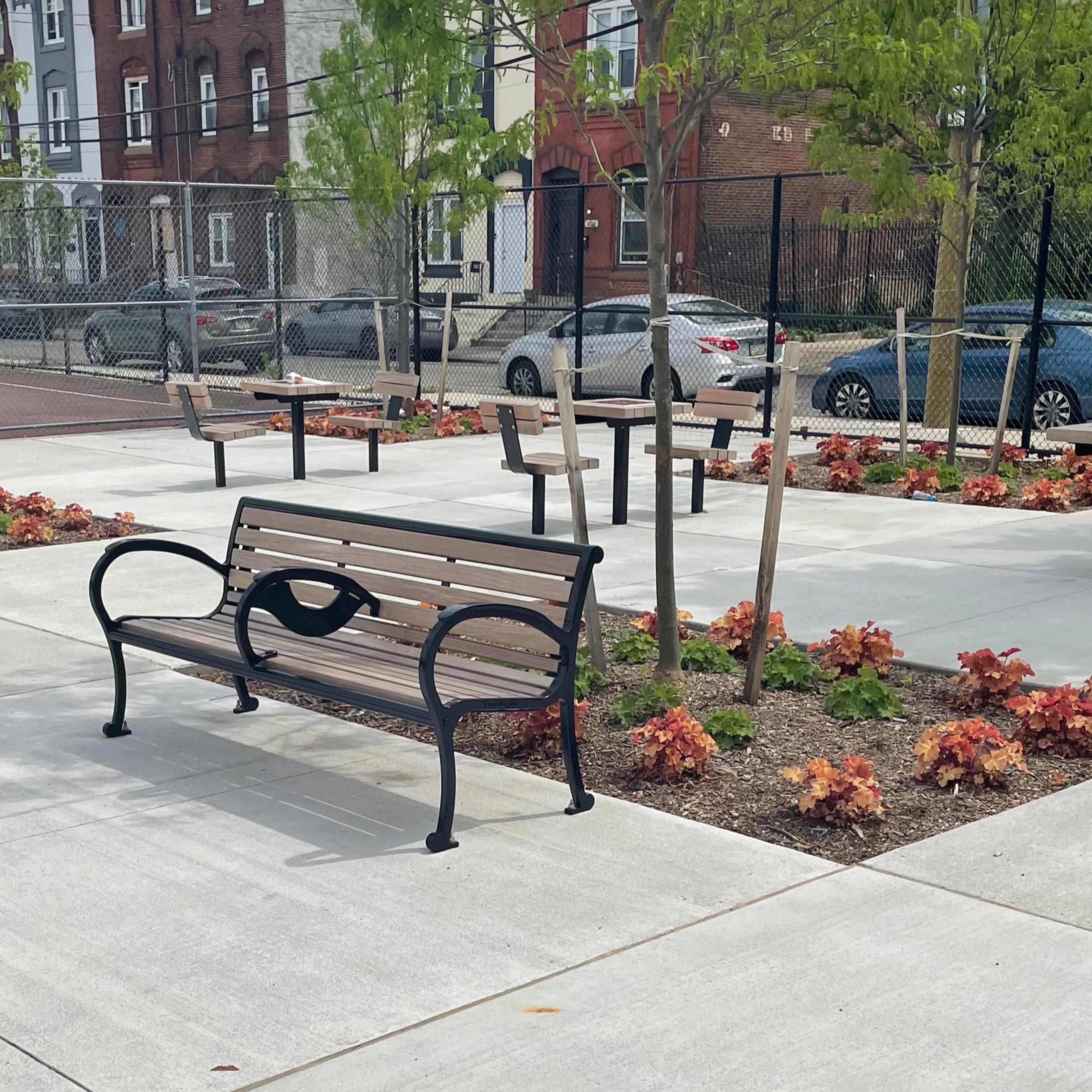 A park area with a bench, several tables, and landscaped flower beds surrounded by a fence.