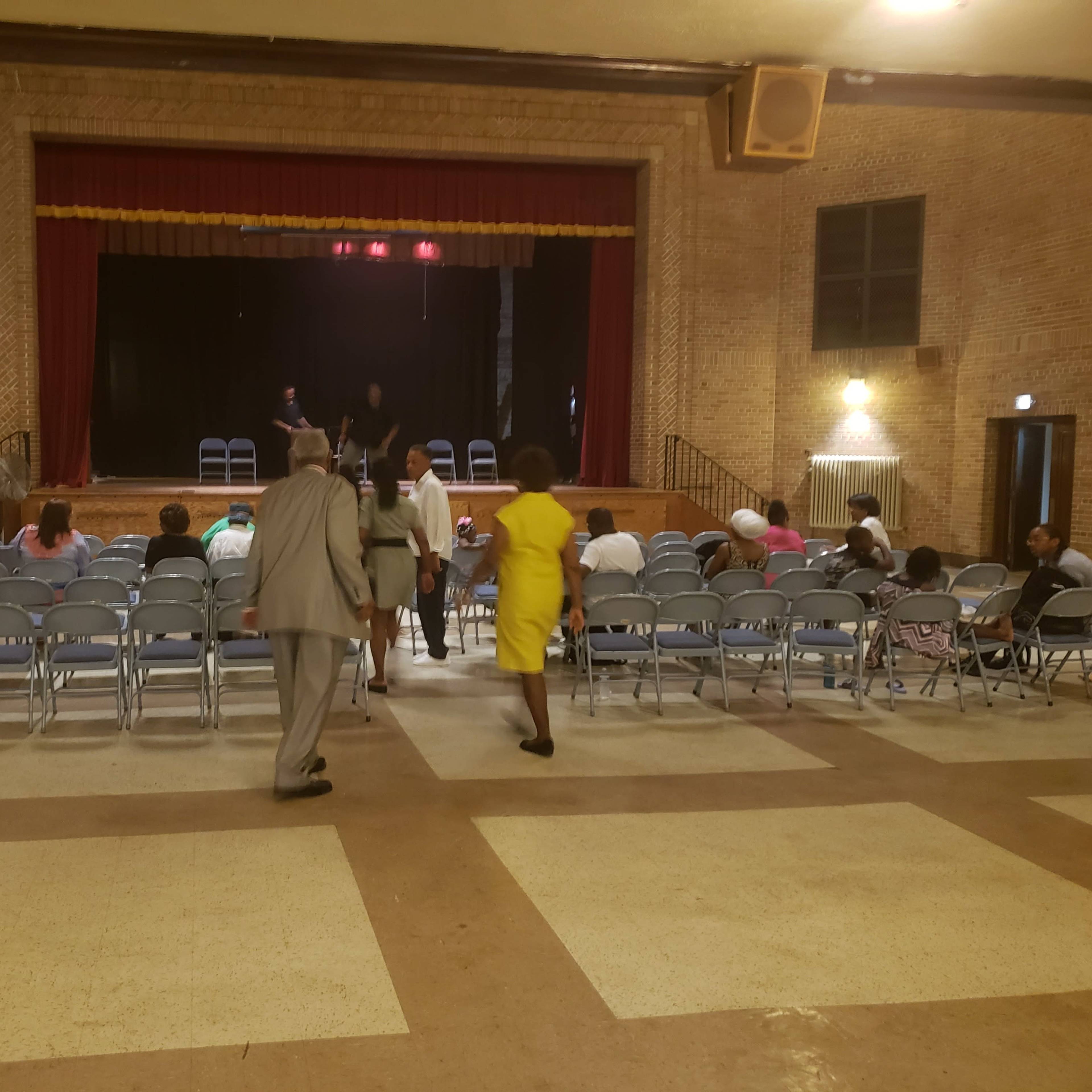 A group of people is entering a performance hall with rows of empty chairs facing a stage.