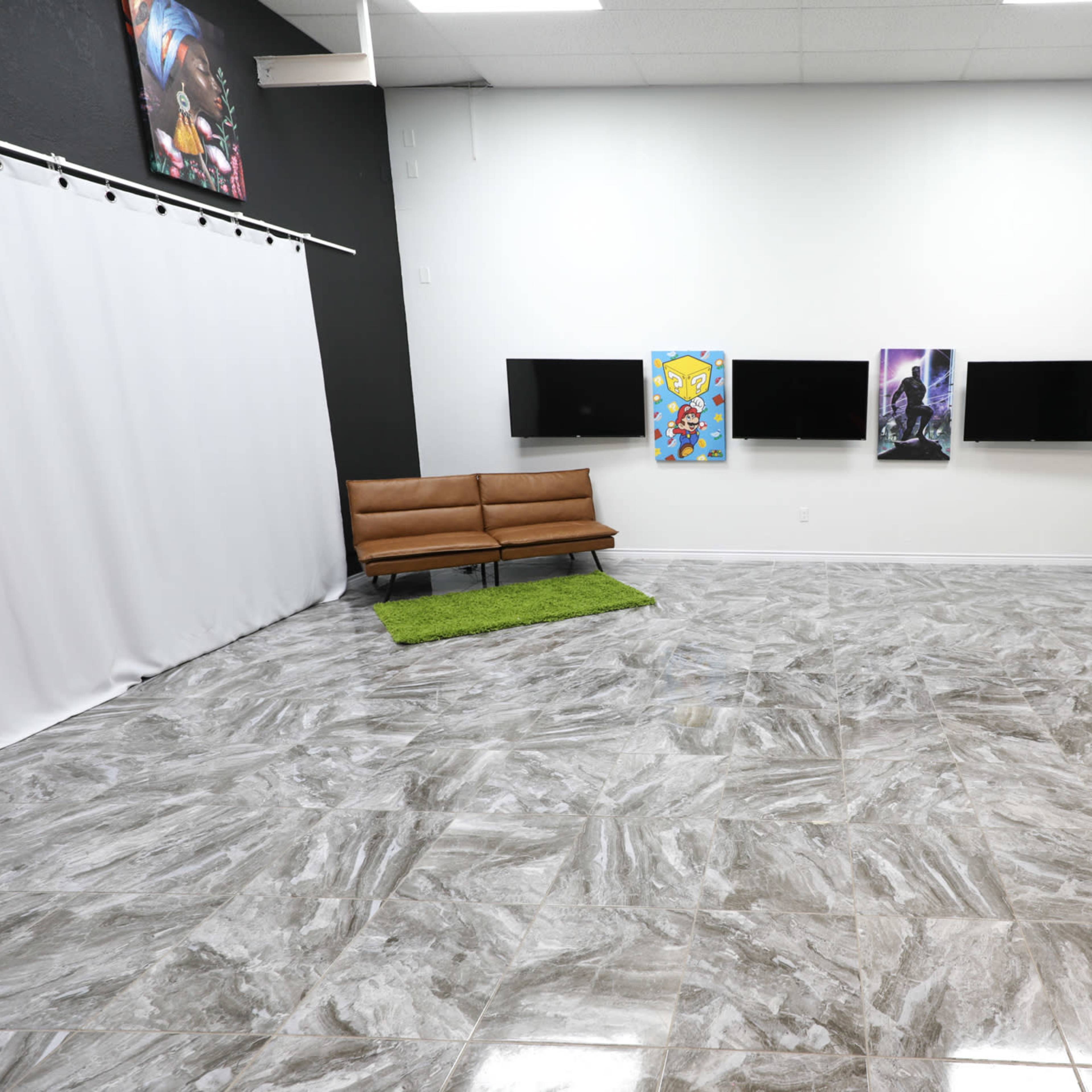 An empty room with gray marble flooring, a brown sofa on a green rug, two television screens mounted on the wall, and artwork displayed on the adjacent wall.