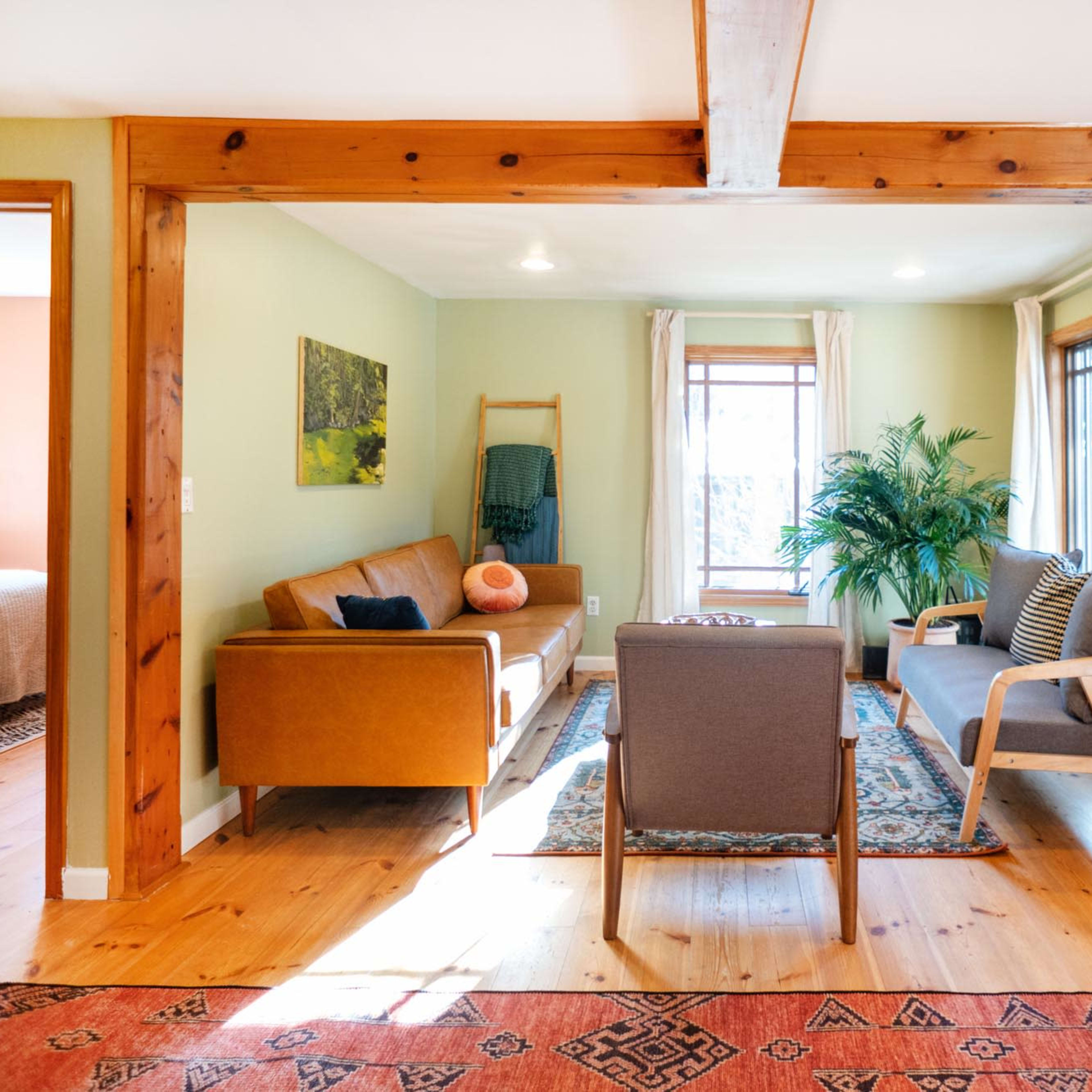 A cozy living room features a brown sofa, a pair of armchairs, a large window, and wooden beams, with light streaming in from outside.