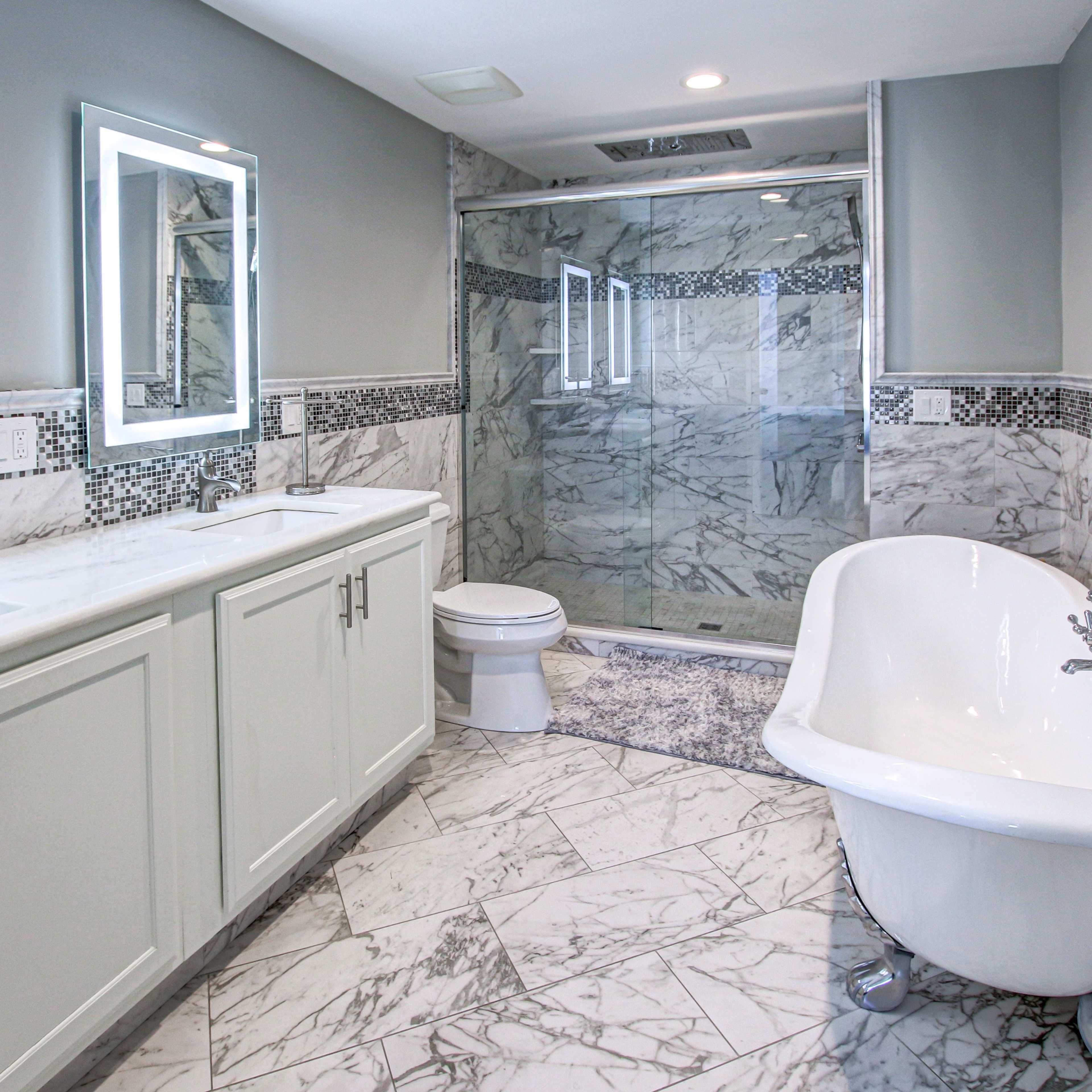 A modern bathroom featuring a marble floor, a freestanding bathtub, double sinks with illuminated mirrors, and a glass shower enclosure.