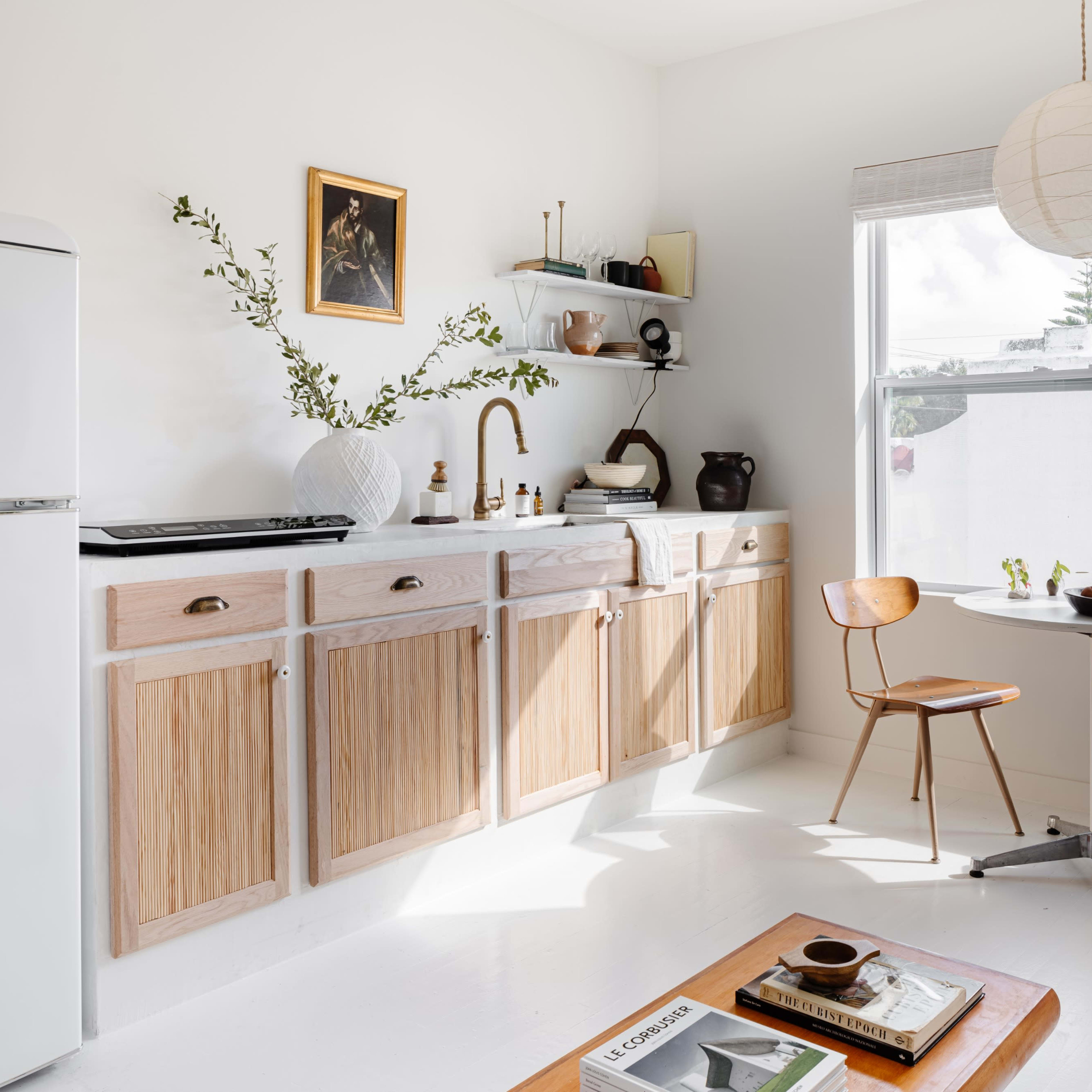 A minimalist kitchen features wooden cabinetry, a vintage refrigerator, and a small dining table with two chairs beside a window.