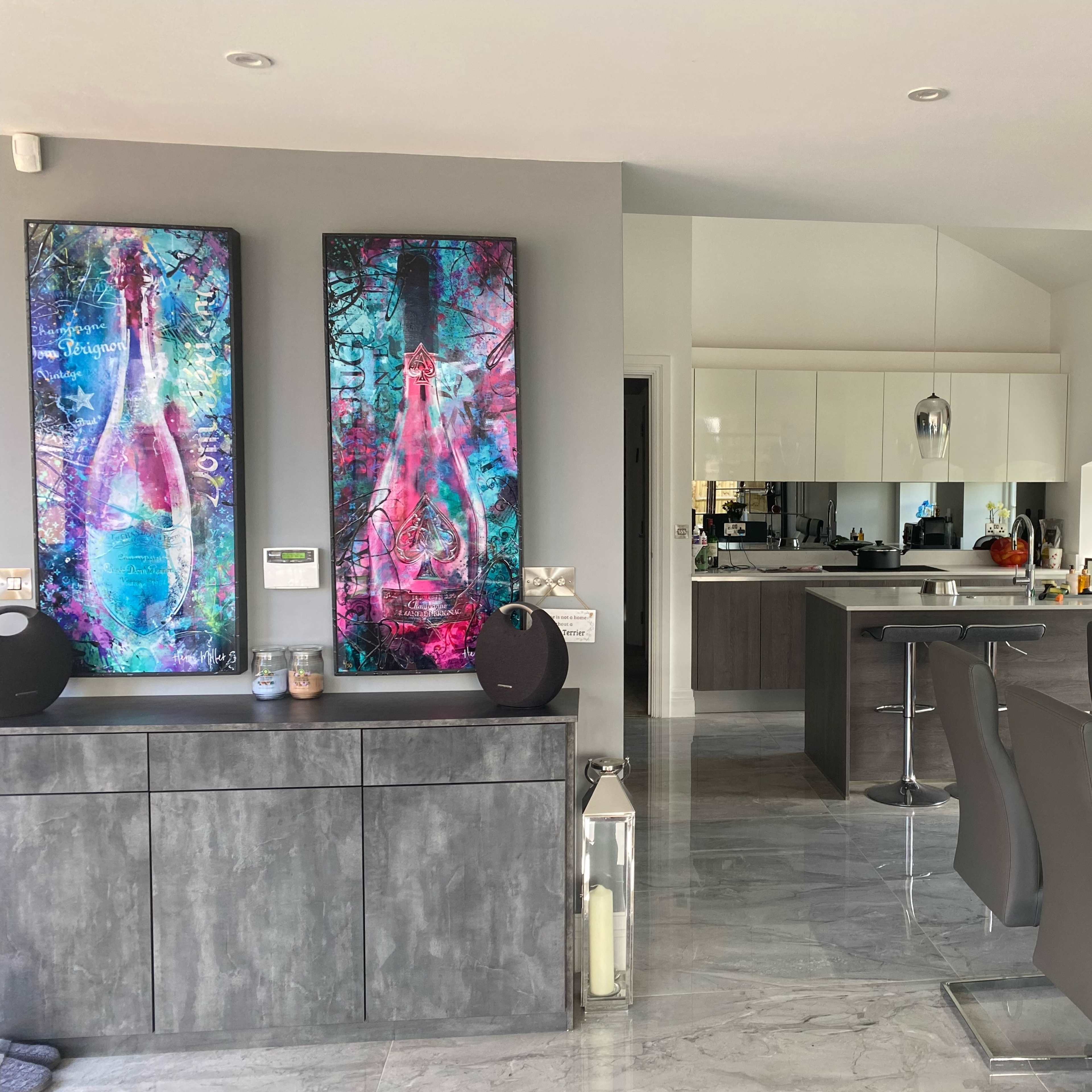 A modern kitchen and dining area featuring gray cabinetry, abstract artwork on the walls, and a glass dining table with gray chairs.