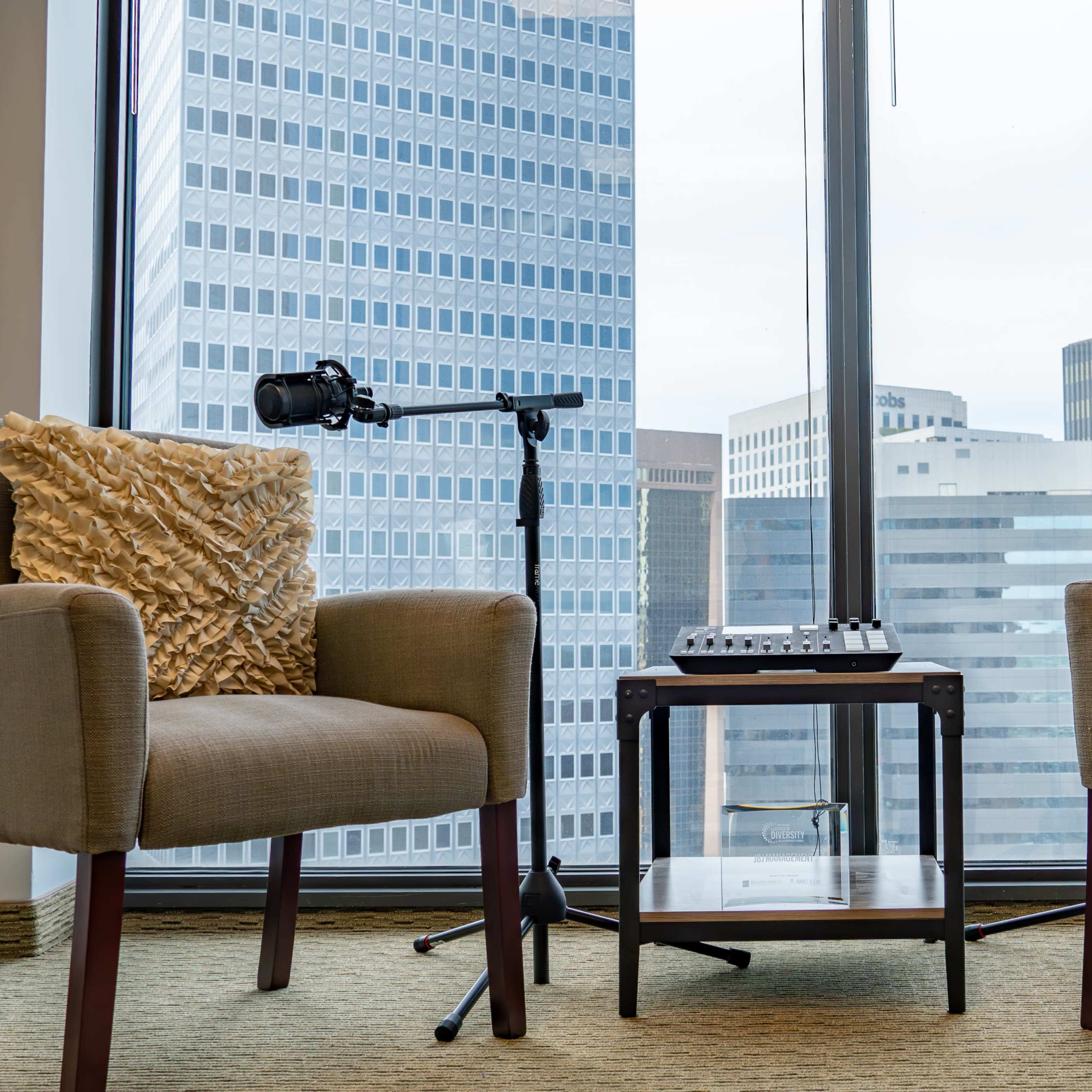 Two upholstered chairs with decorative pillows are positioned near a window, with a small table and microphone stands between them, overlooking a city skyline.