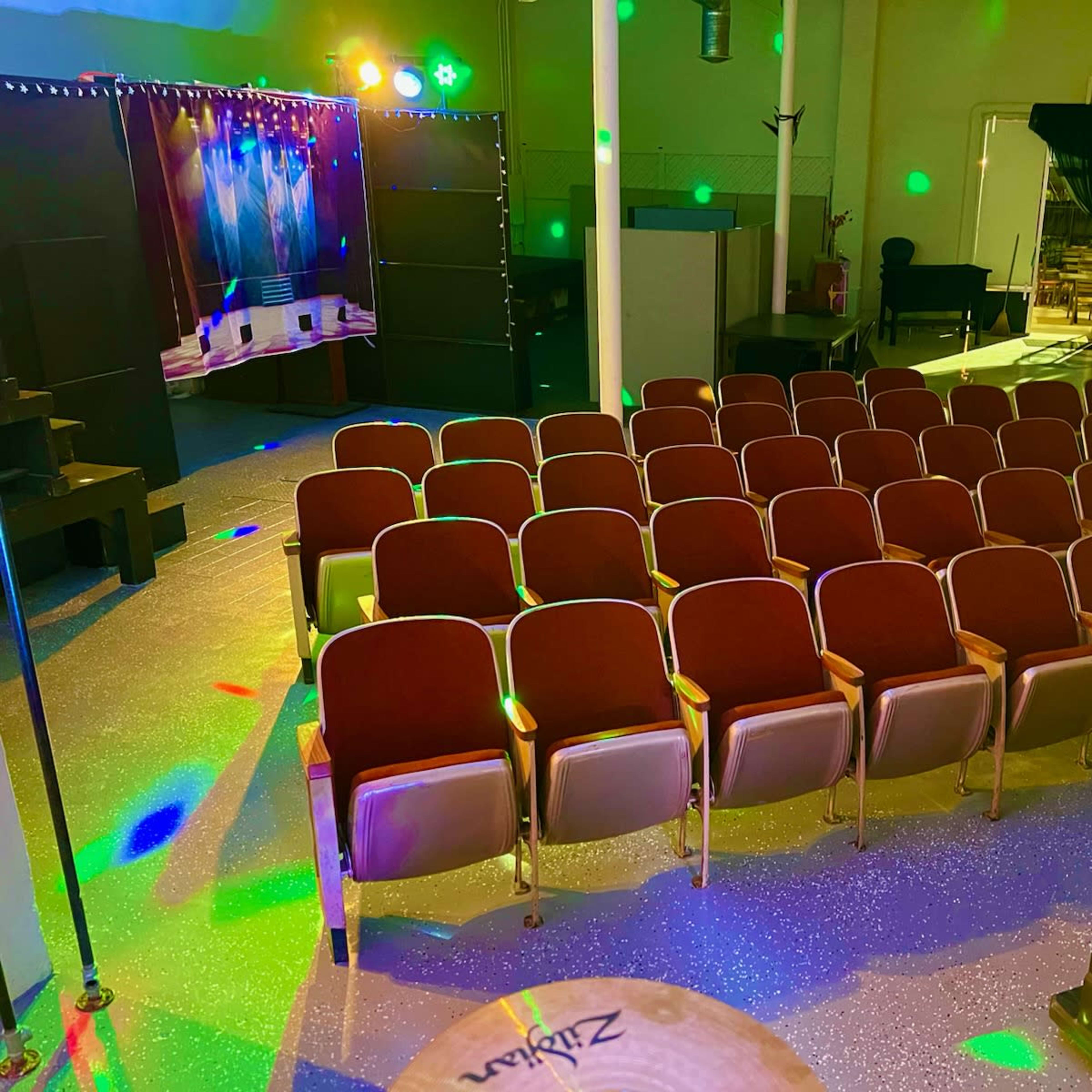 An empty auditorium with rows of maroon seats arranged in front of a stage, illuminated by colorful lights.