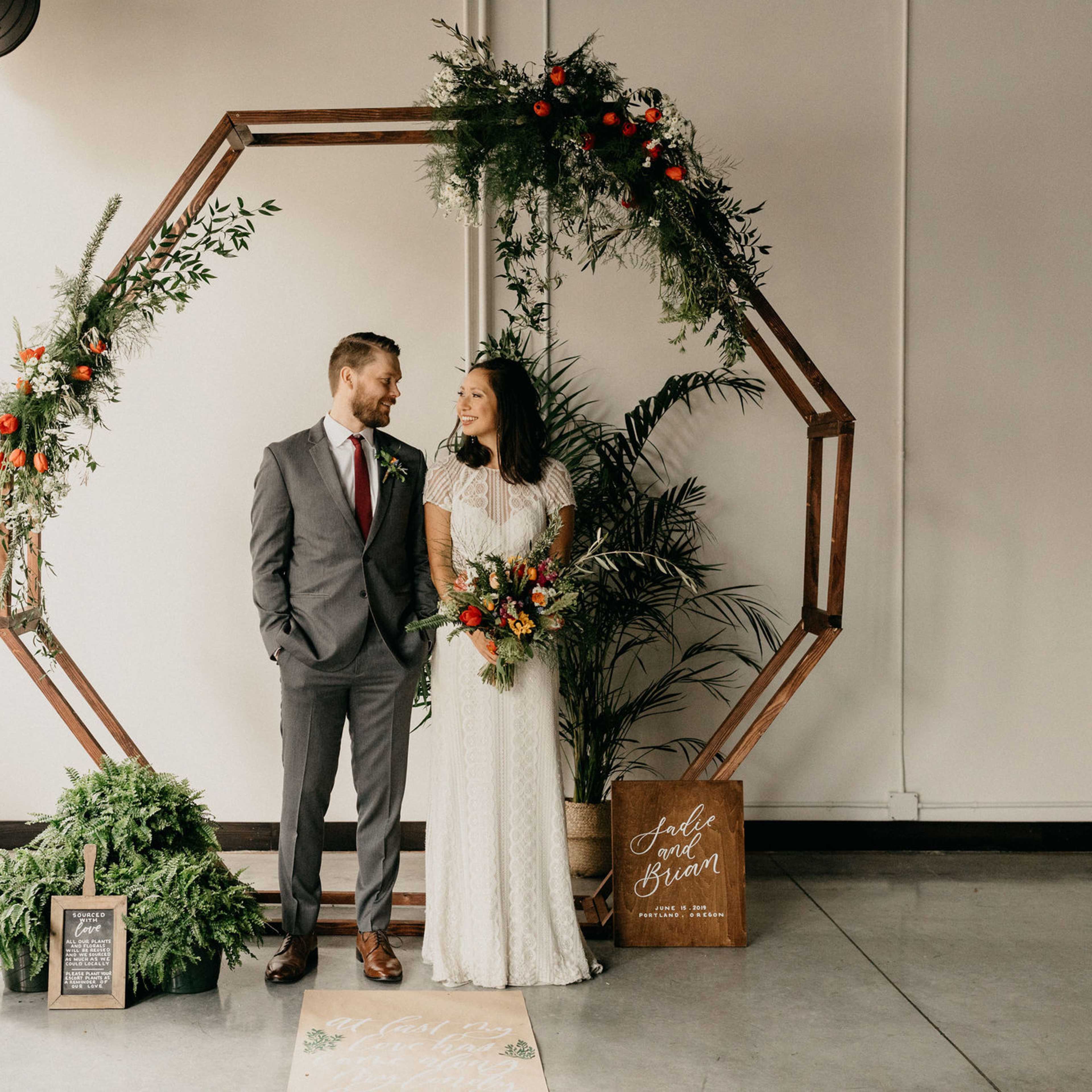 A couple stands together in front of a wooden octagonal arch adorned with greenery and orange accents, while a sign nearby displays a decorative message.