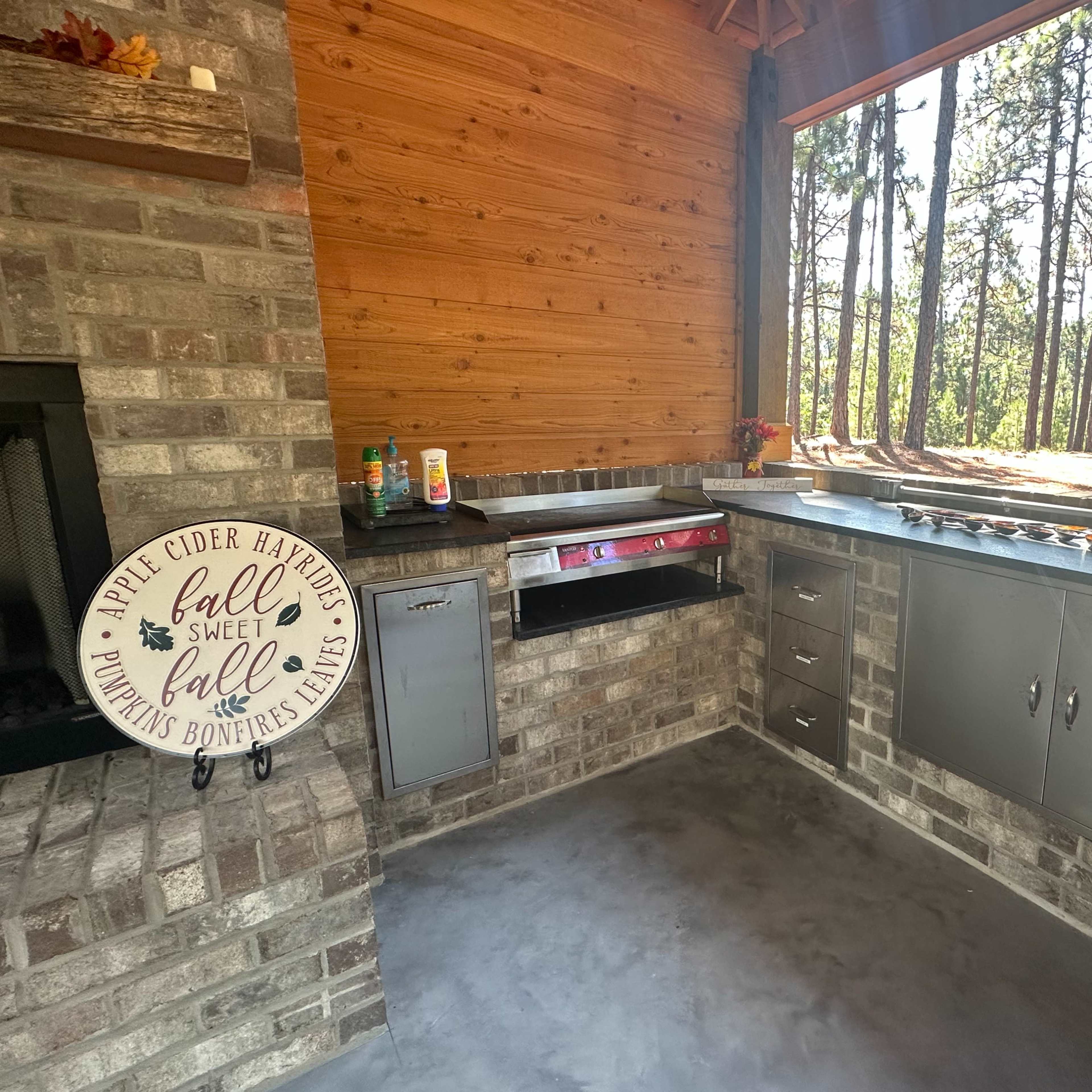 A spacious outdoor kitchen area with a stone wall, stainless steel appliances, and a wooden ceiling, surrounded by trees.