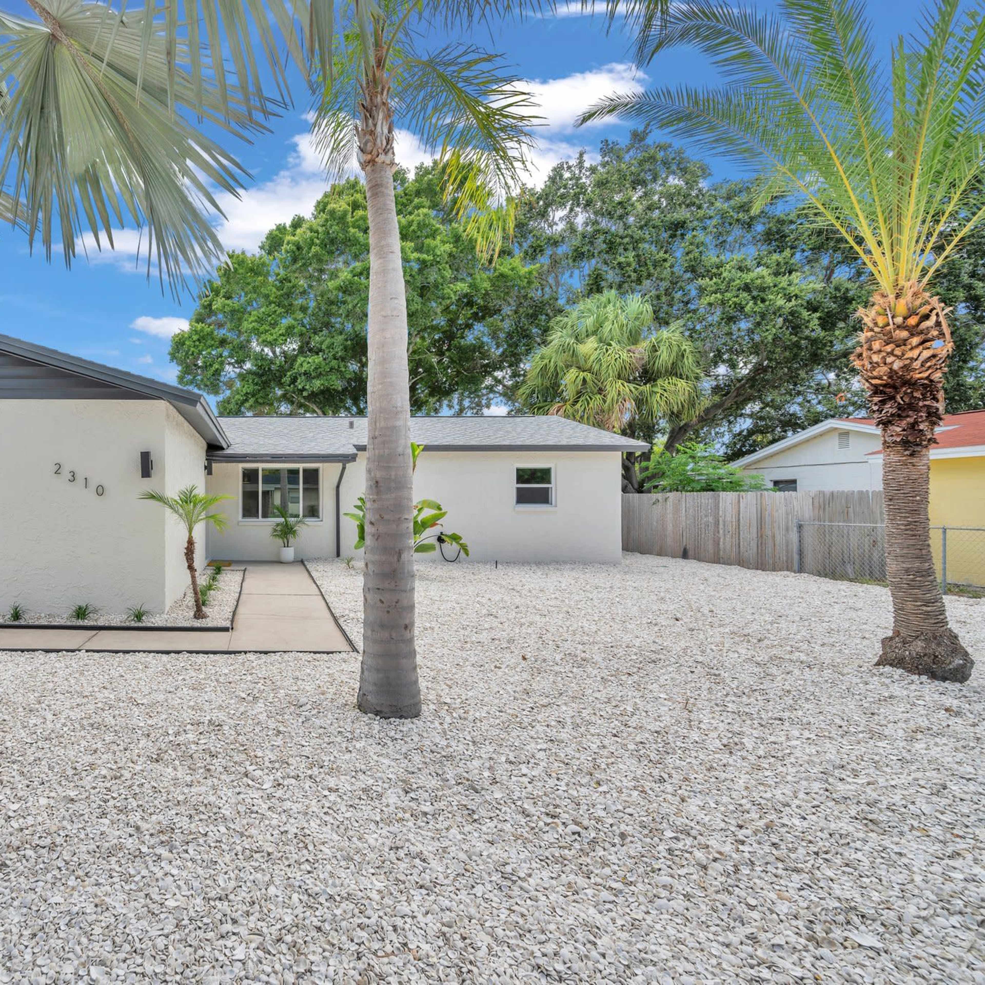 A single-story house with a garage, surrounded by a gravel yard and palm trees.