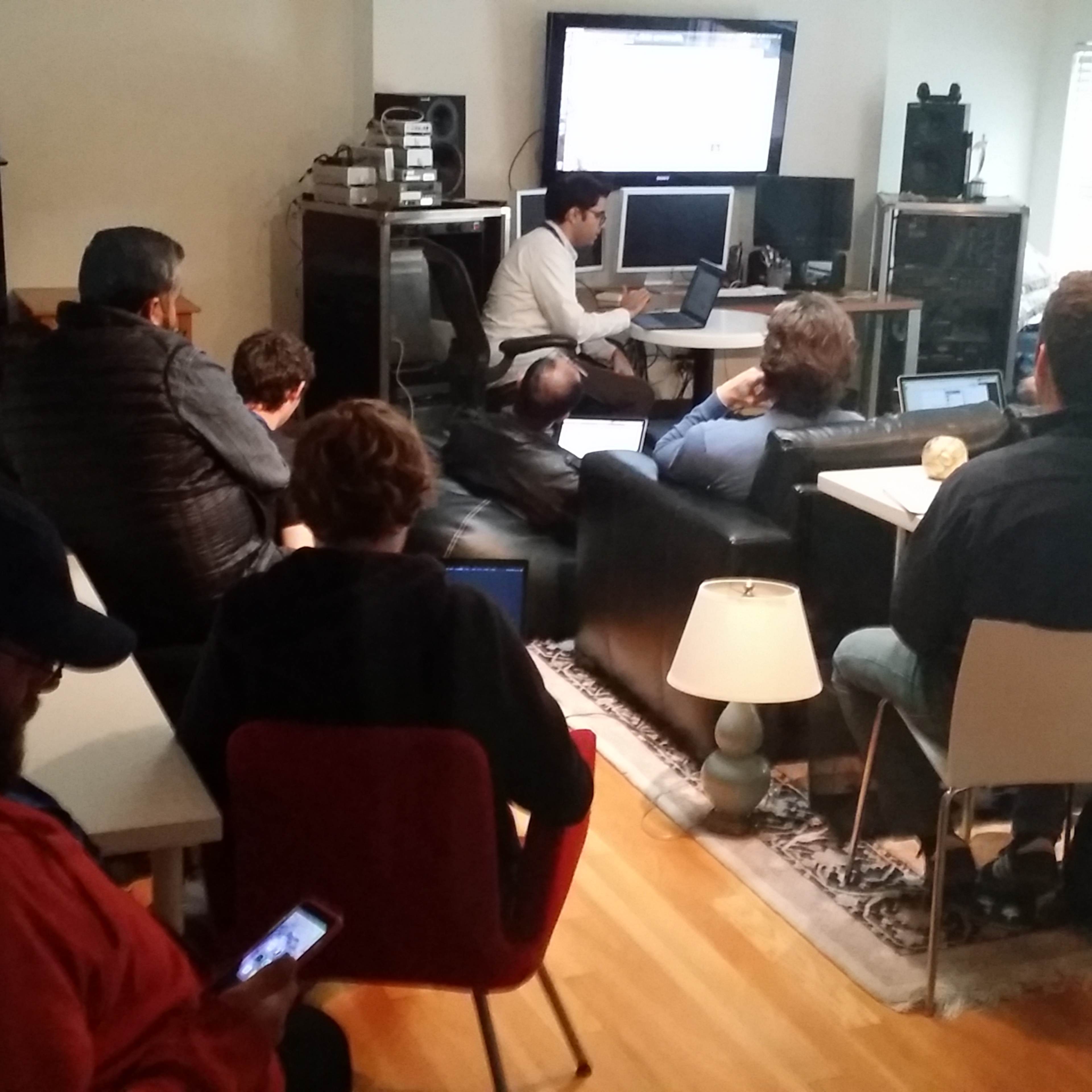 A group of people is seated in a living room, attentively watching a presentation on a screen while some use laptops and smartphones.