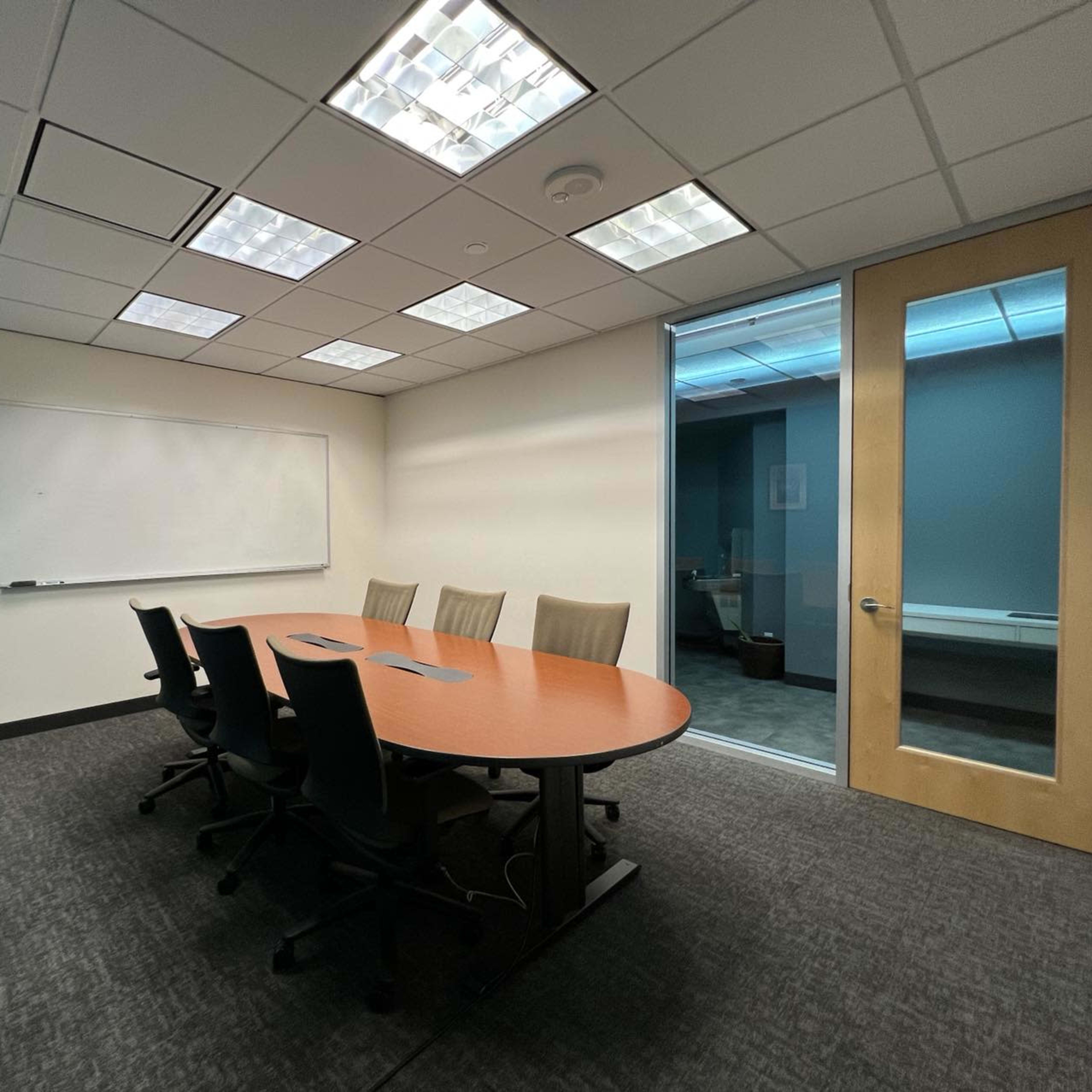 A conference room features a wooden table surrounded by chairs, a whiteboard on the wall, and a glass door leading to another area.