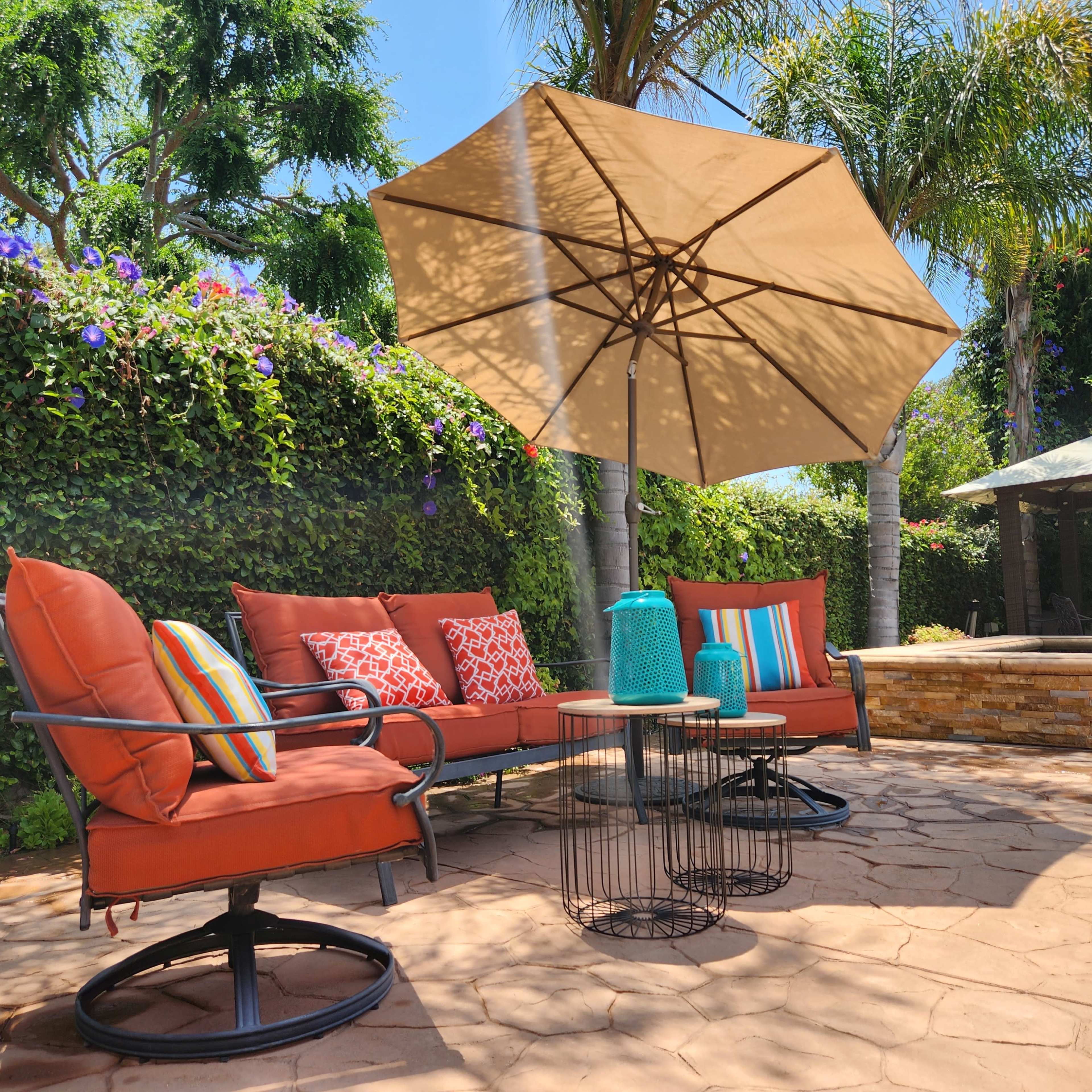 A patio area features a seating arrangement with orange cushions, a round table, and an umbrella, surrounded by greenery and flowering plants.
