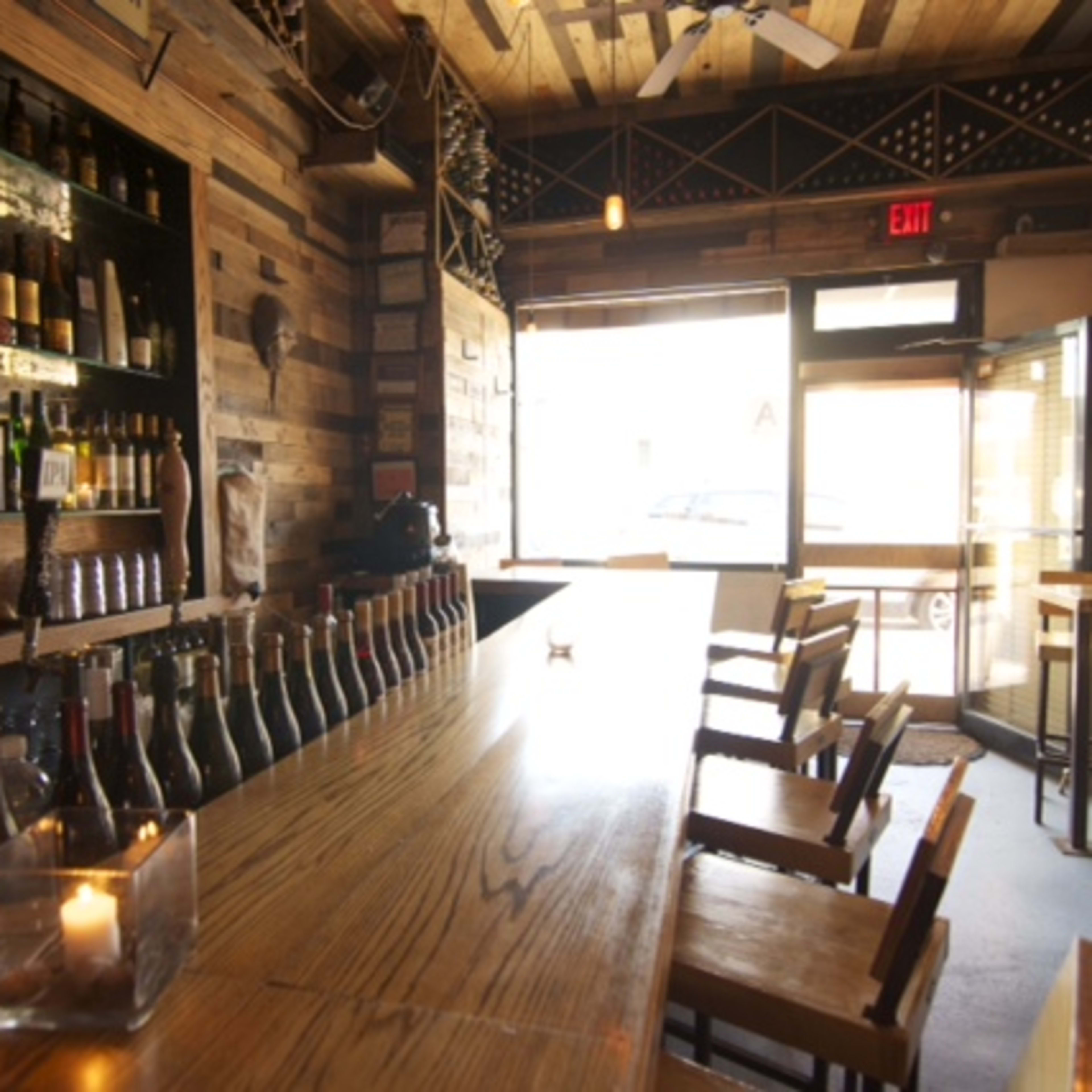 The interior of a restaurant or bar with a wooden counter, shelves of wine bottles, and a seating area with wooden stools and tables.