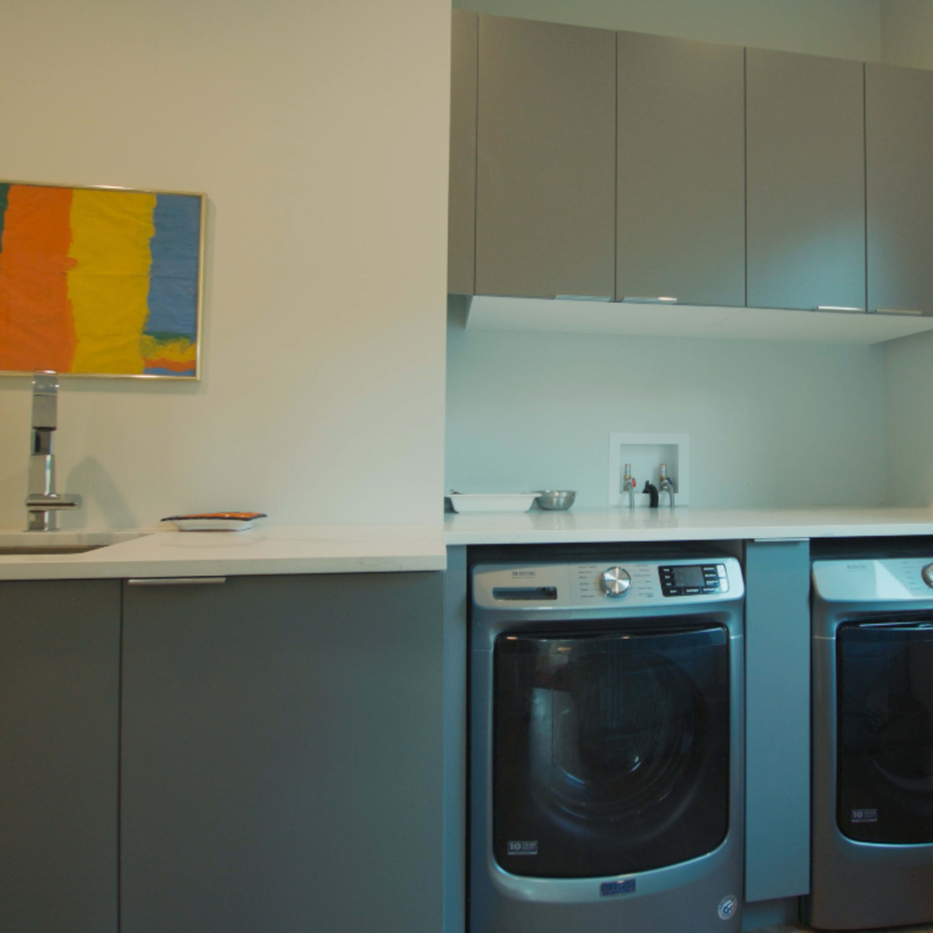 A laundry room features stacked silver washing and drying appliances beneath a countertop, alongside gray cabinets and a colorful abstract painting on the wall.