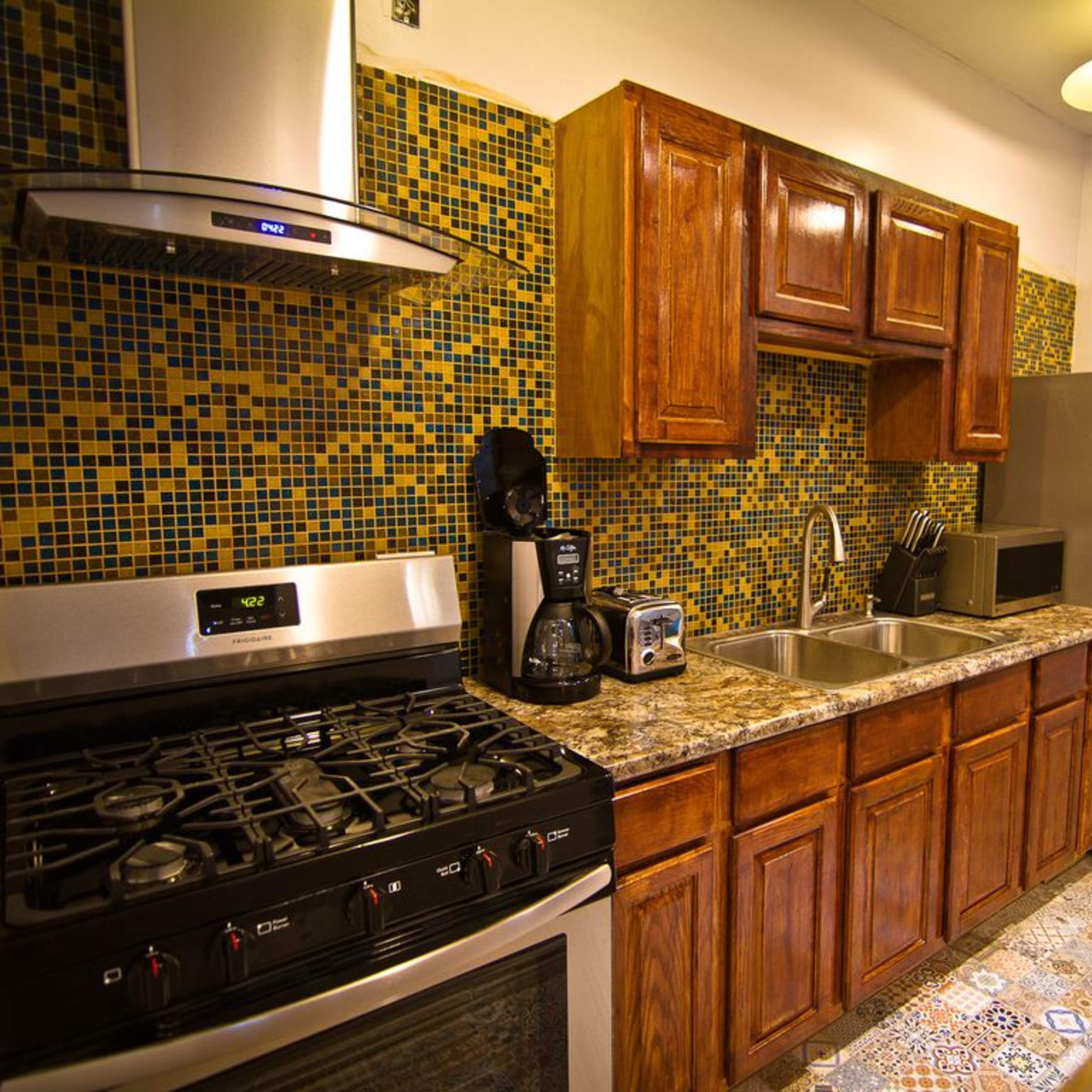 A kitchen with wooden cabinets, a stainless steel stove, a double sink, and a refrigerator, featuring a colorful tiled backsplash.