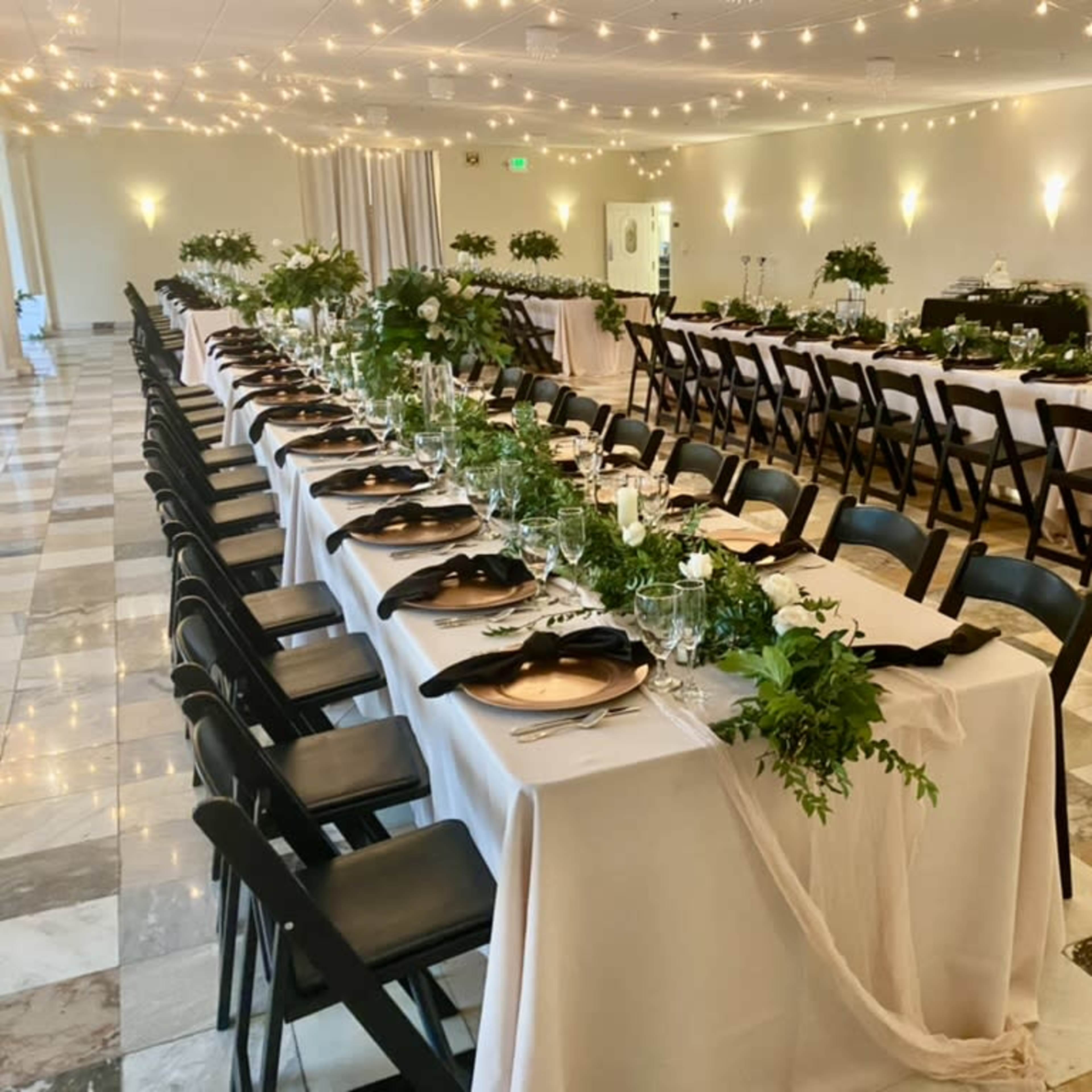 A large dining area is set with multiple long tables decorated with white tablecloths, black napkins, and greenery, illuminated by string lights overhead.