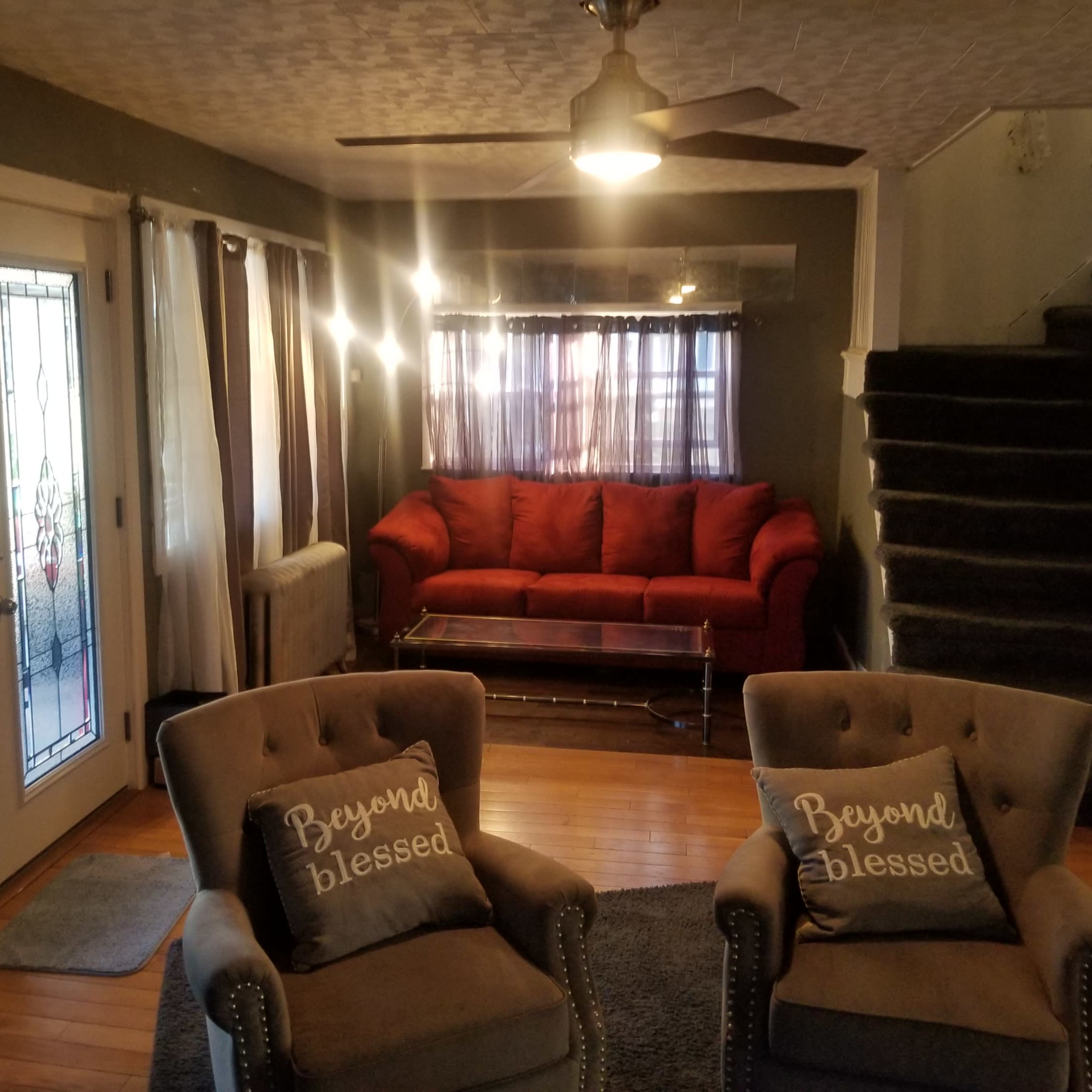 A living room with a red sofa, two gray armchairs with decorative cushions, a wooden coffee table, and a staircase in the background.