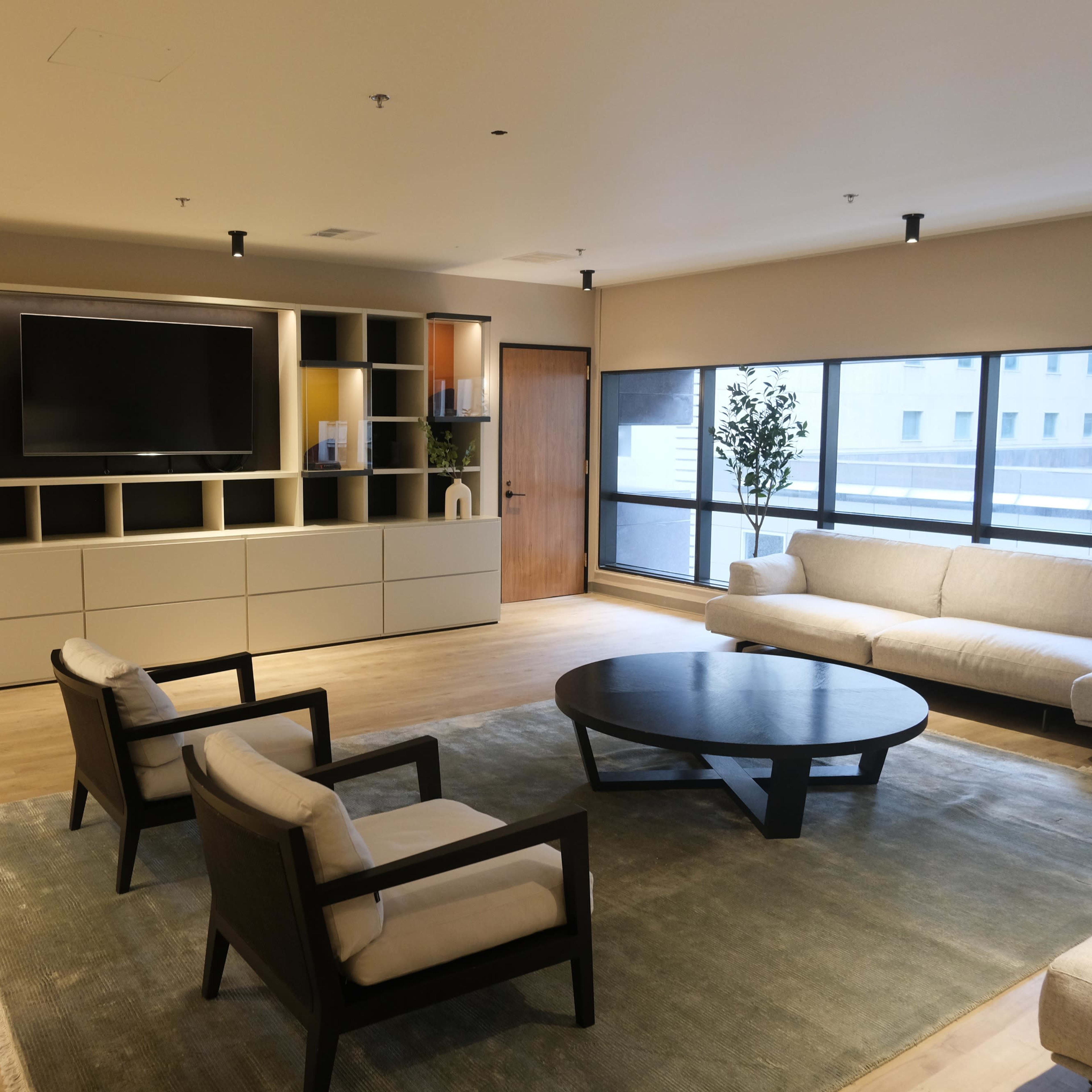 A modern living room with a large TV mounted on a wall unit, a round coffee table, two armchairs, and a sofa facing large windows.