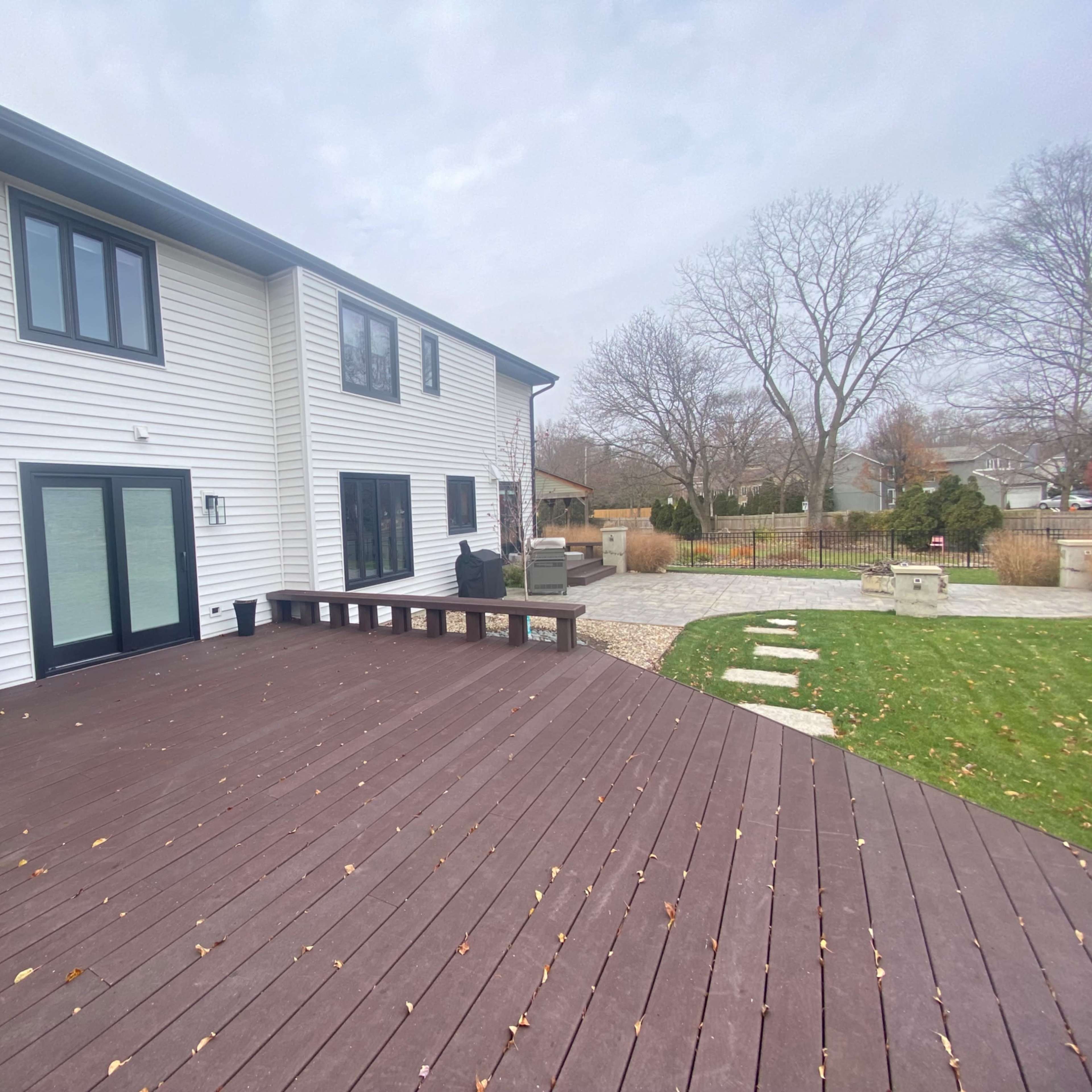 A modern two-story house with white siding and large windows, accompanied by a wooden deck and a landscaped yard featuring stone pathways and grass.