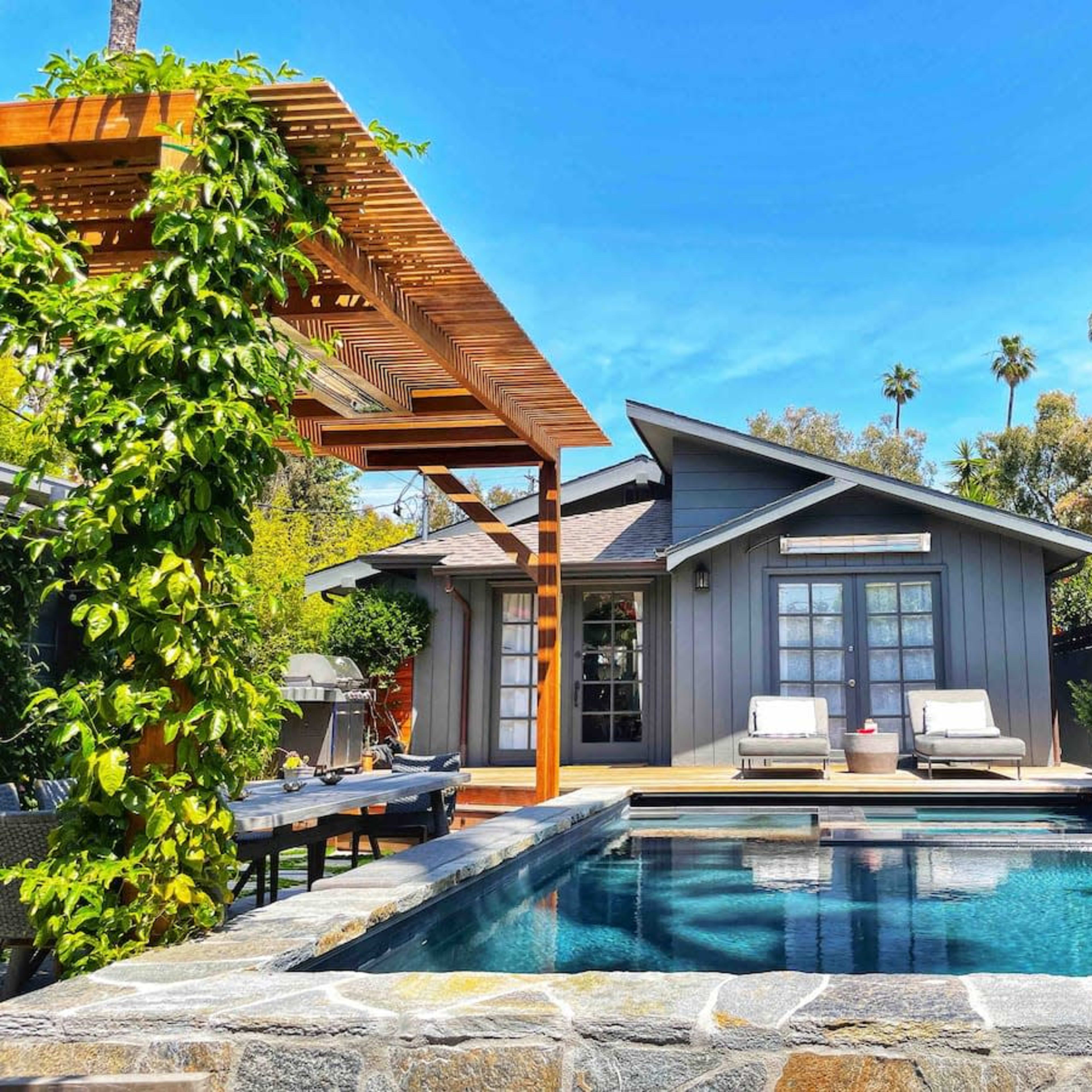 A modern house with a gray exterior sits beside a swimming pool surrounded by greenery and patio furniture.