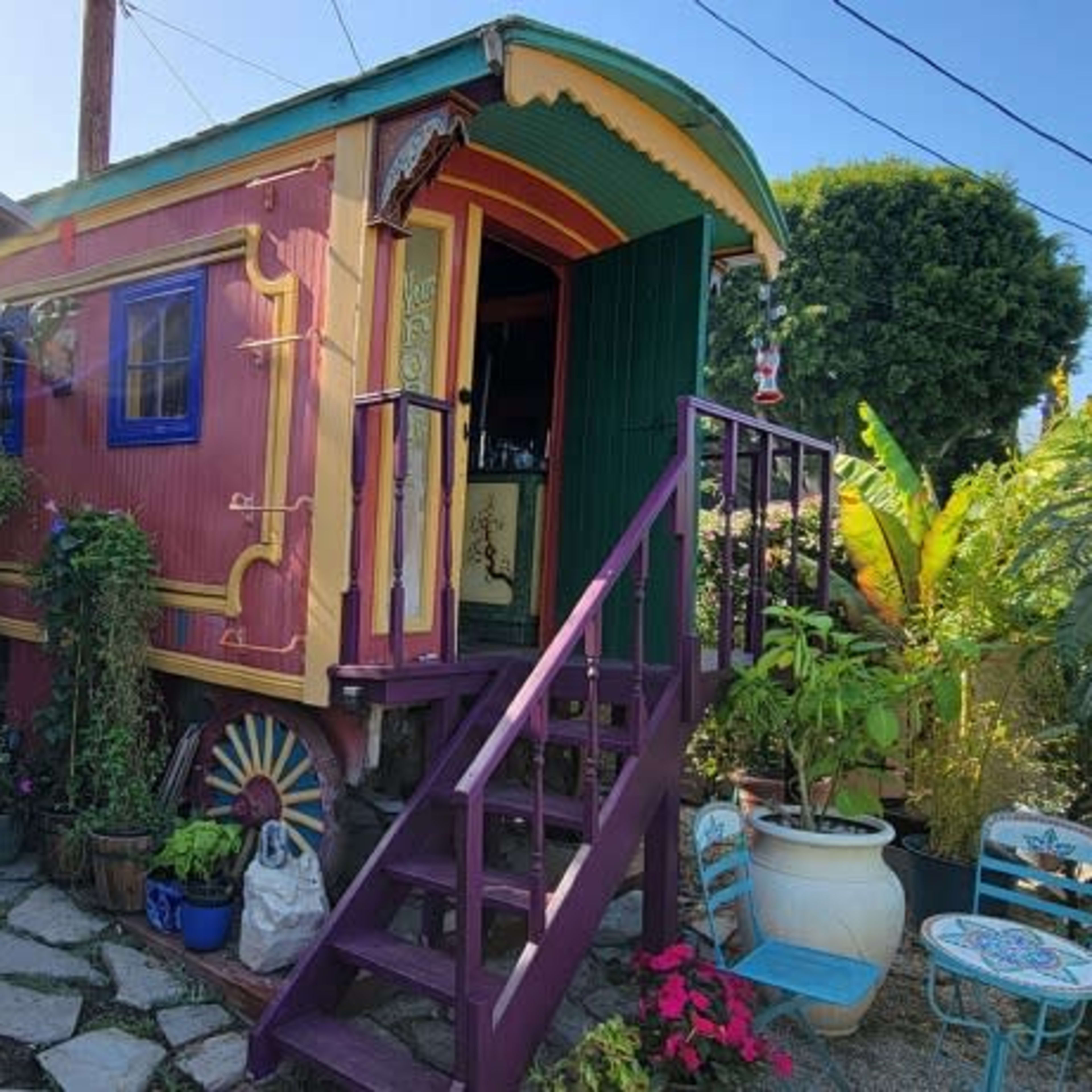 A colorful caravan with a decorated exterior is positioned on a stone path surrounded by various plants and pots.