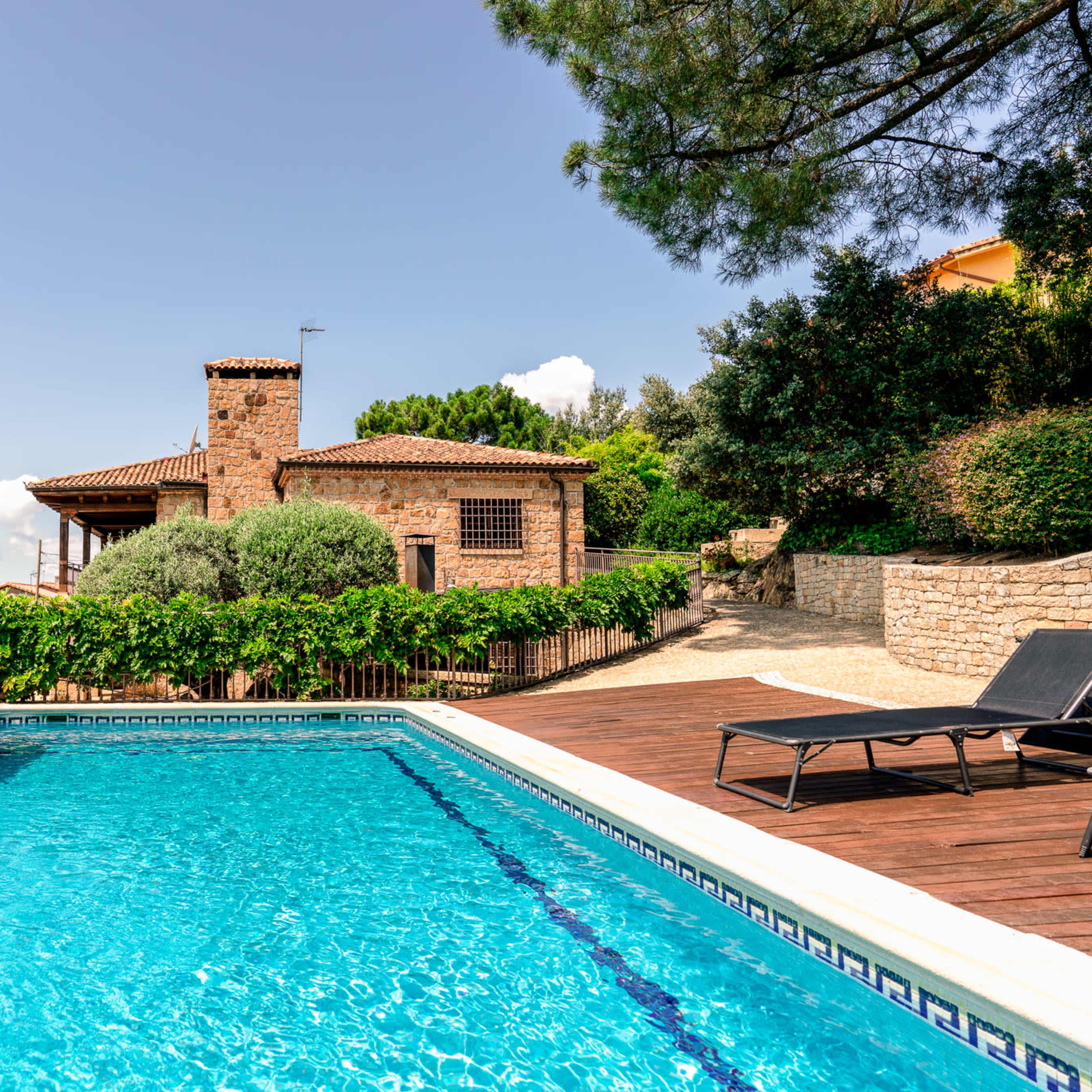 A stone house with a pool is surrounded by lush greenery and wooden decking under a clear sky.