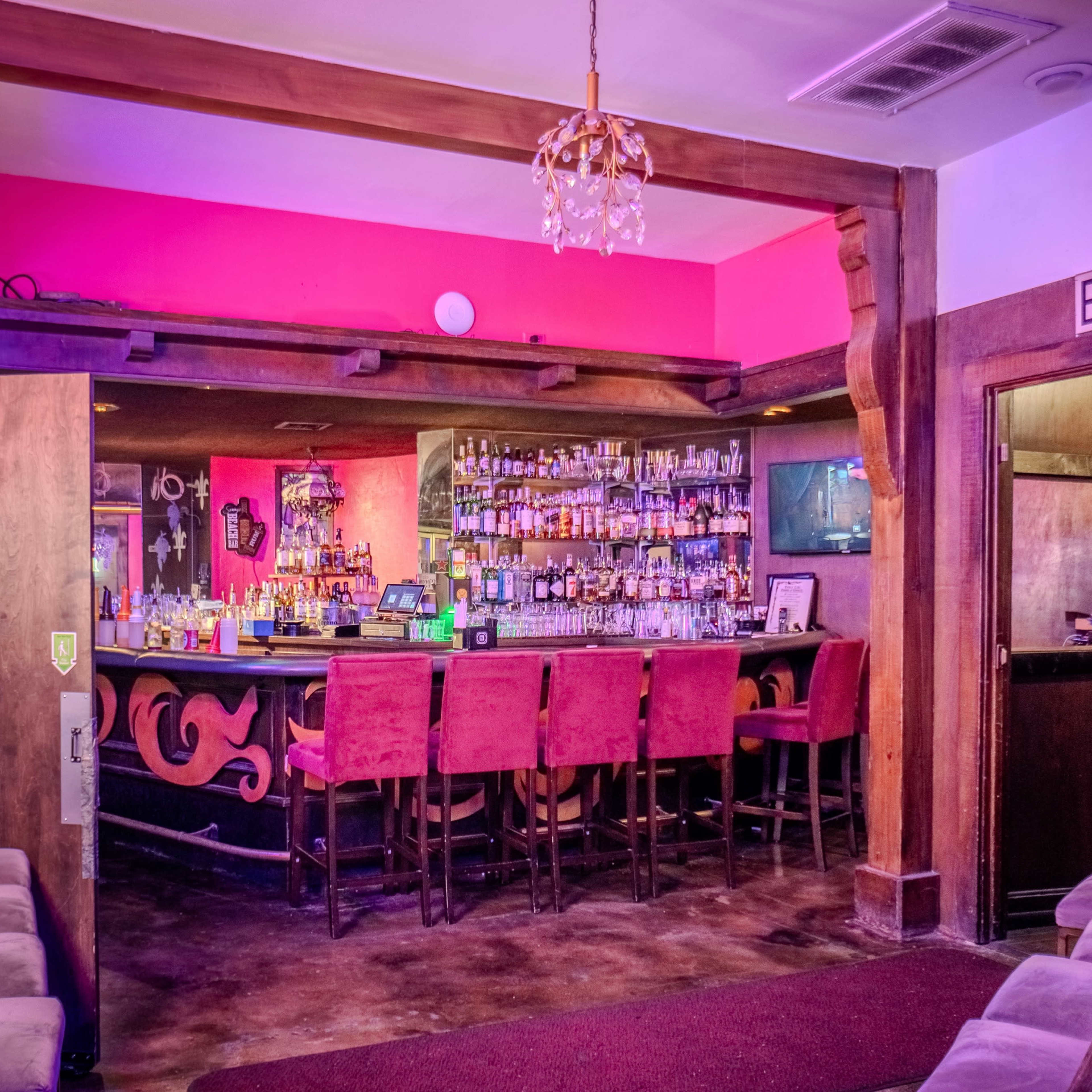A dimly lit bar area with a long counter lined with high stools, shelves stacked with various bottles of alcohol, and neon signage illuminating the walls.
