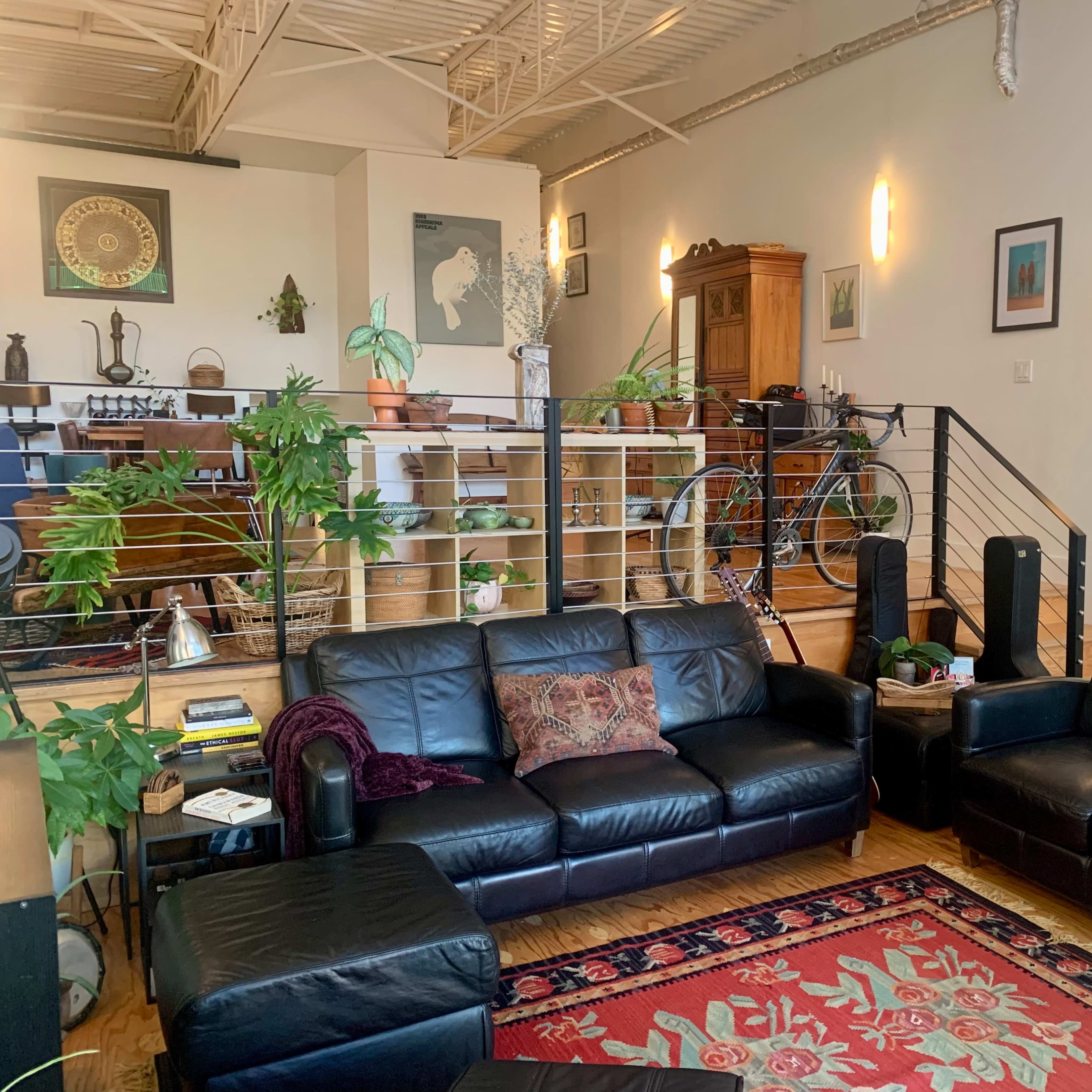 A modern living room with black leather sofas, a coffee table, and a variety of plants, leading to an elevated dining area with wooden furniture and a bicycle in the background.