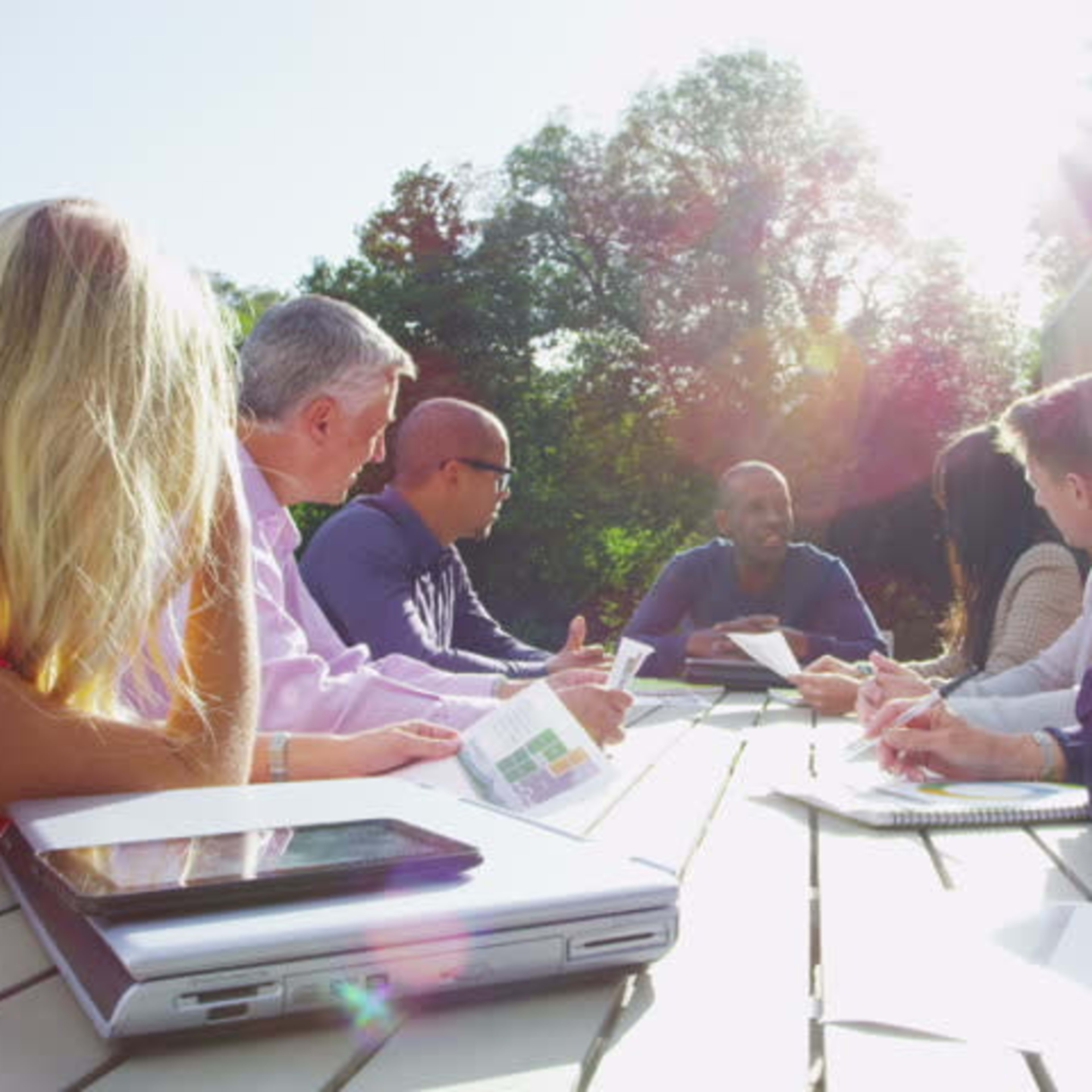 A diverse group of professionals is seated around a table outdoors, engaging in a discussion while reviewing documents.