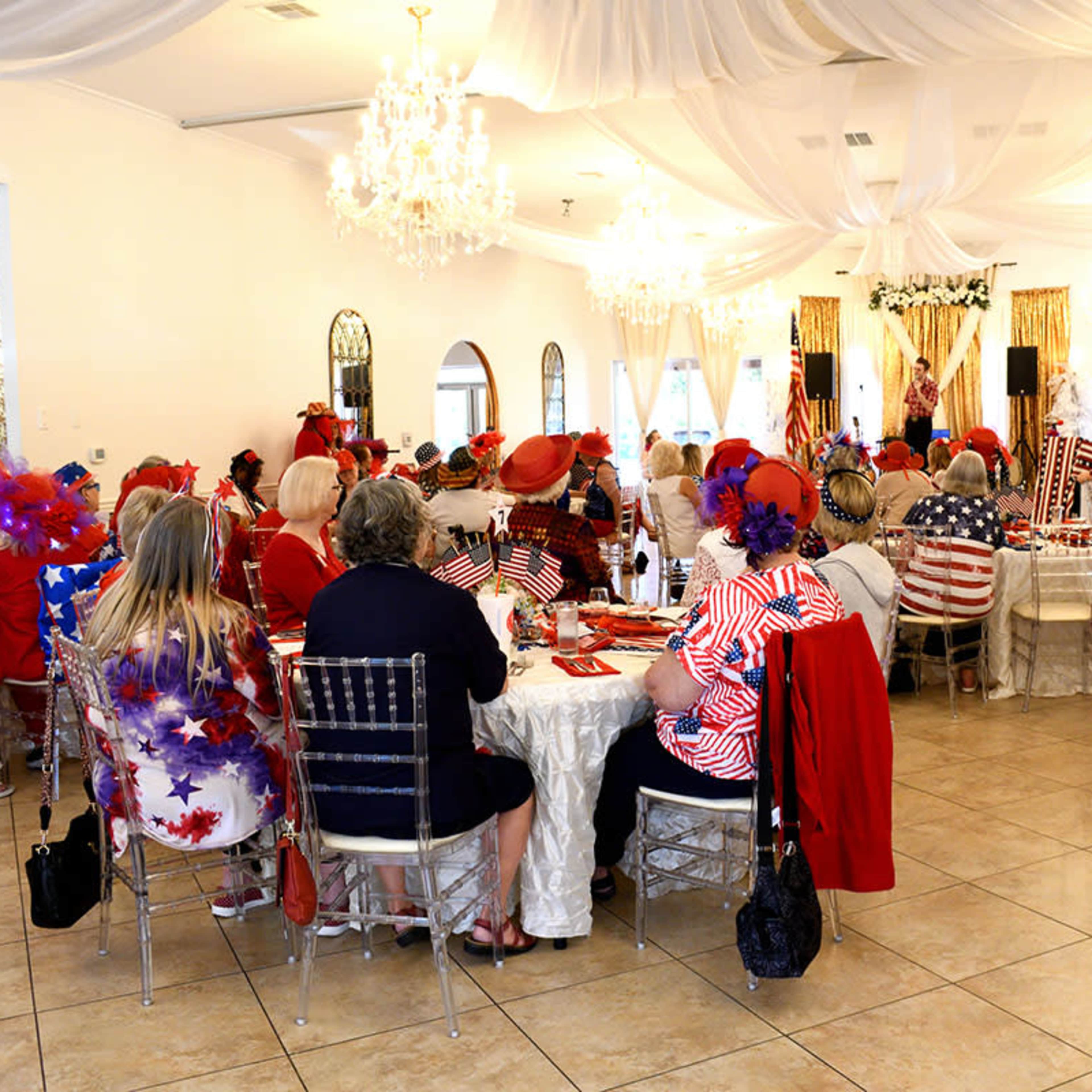 A large group of people in festive attire are seated around tables in a banquet hall decorated with chandeliers and draped fabric.