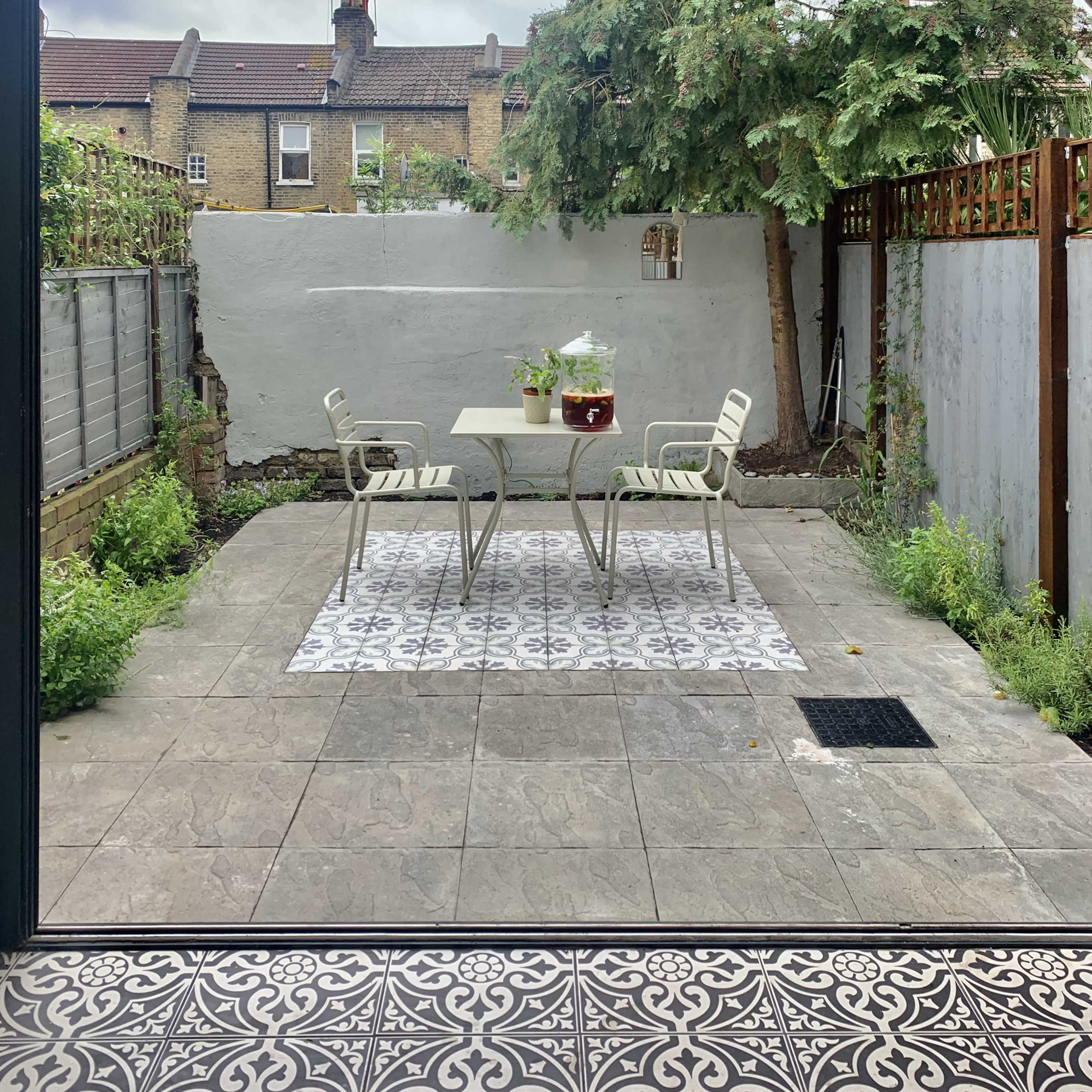 A small outdoor courtyard with a tiled floor, featuring a table and chairs on a patterned rug, surrounded by walls and greenery.
