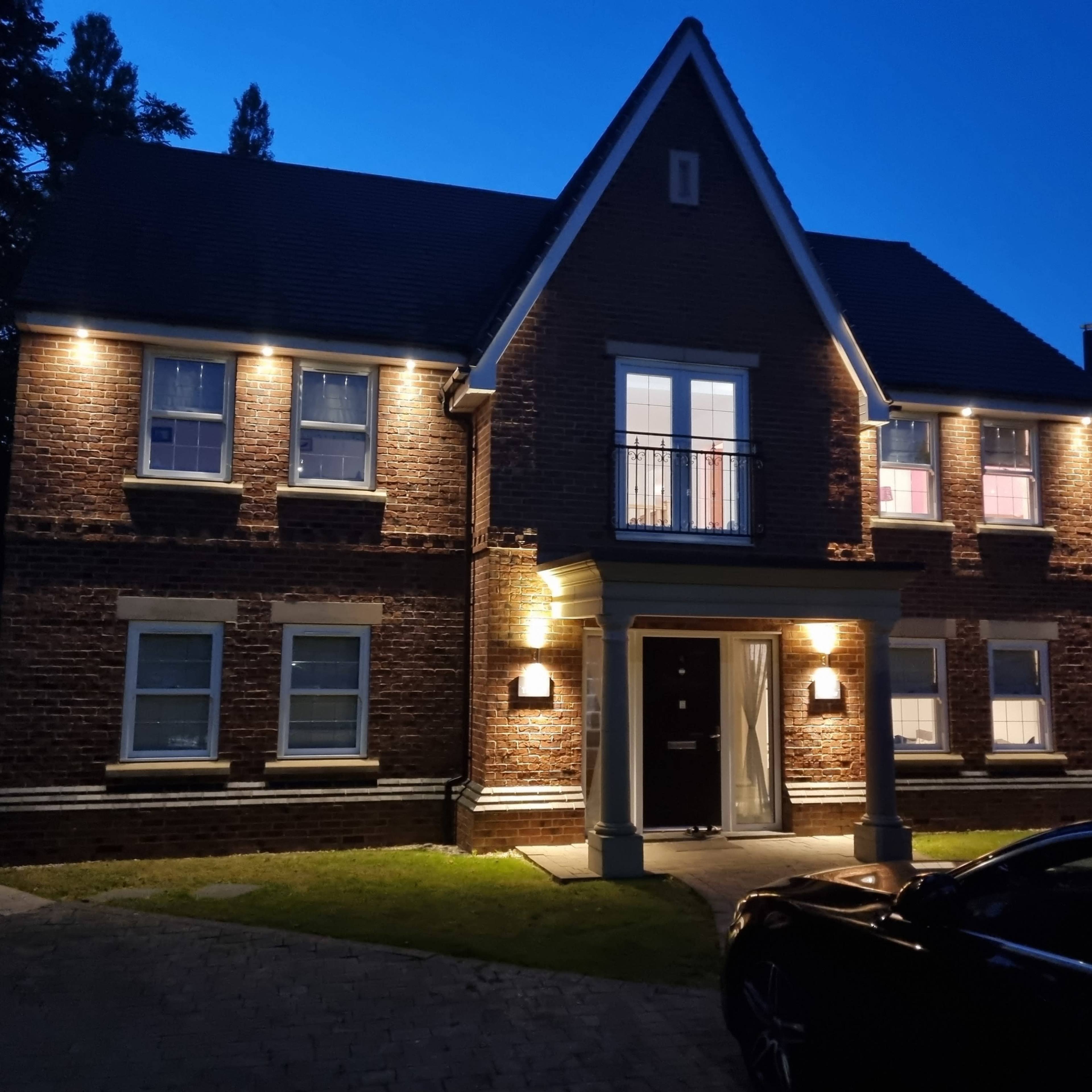A two-story brick house is illuminated by lights at dusk, featuring multiple windows and a central front door.