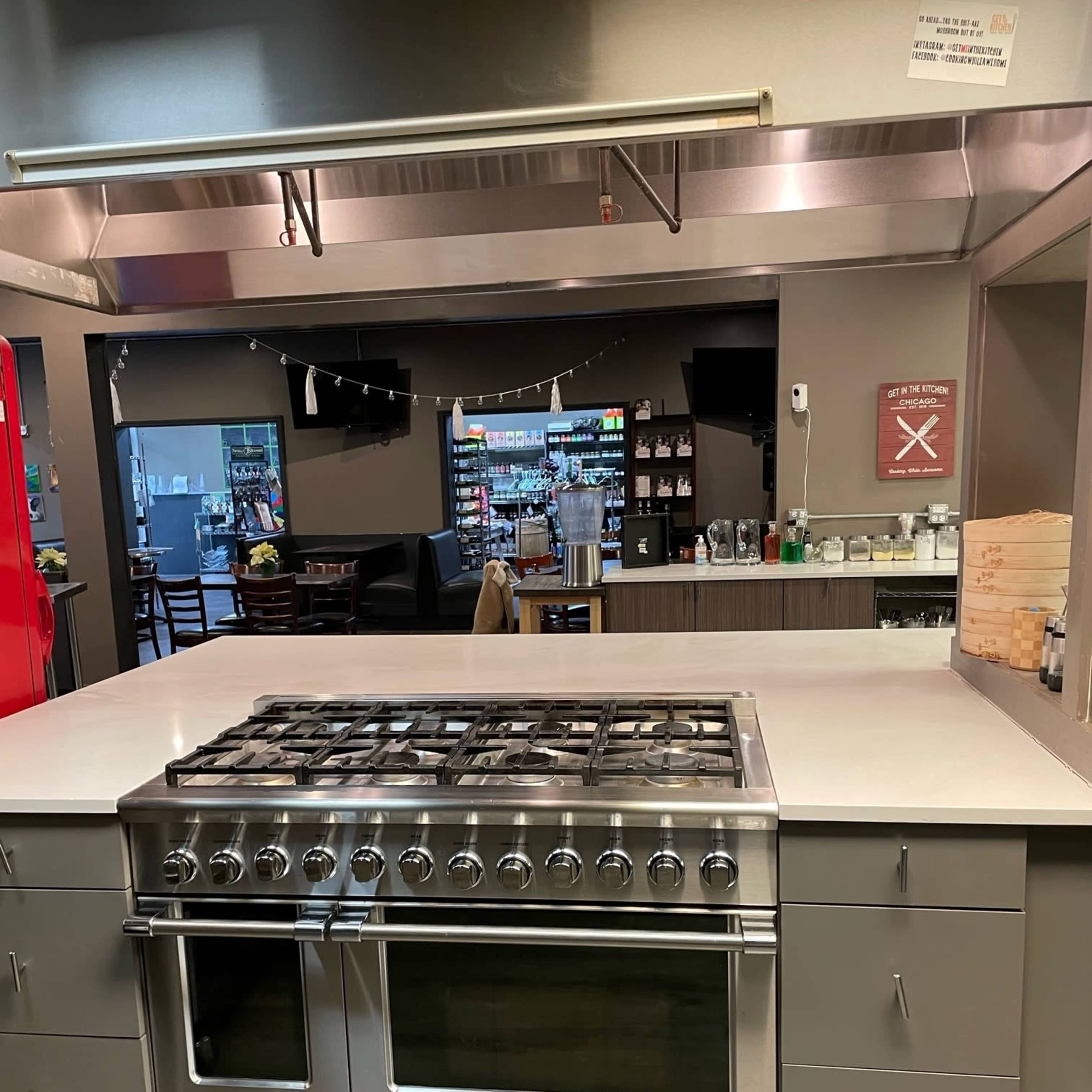 A modern kitchen with a large stainless steel gas range on an island countertop and a background featuring a dining area and shelves of supplies.