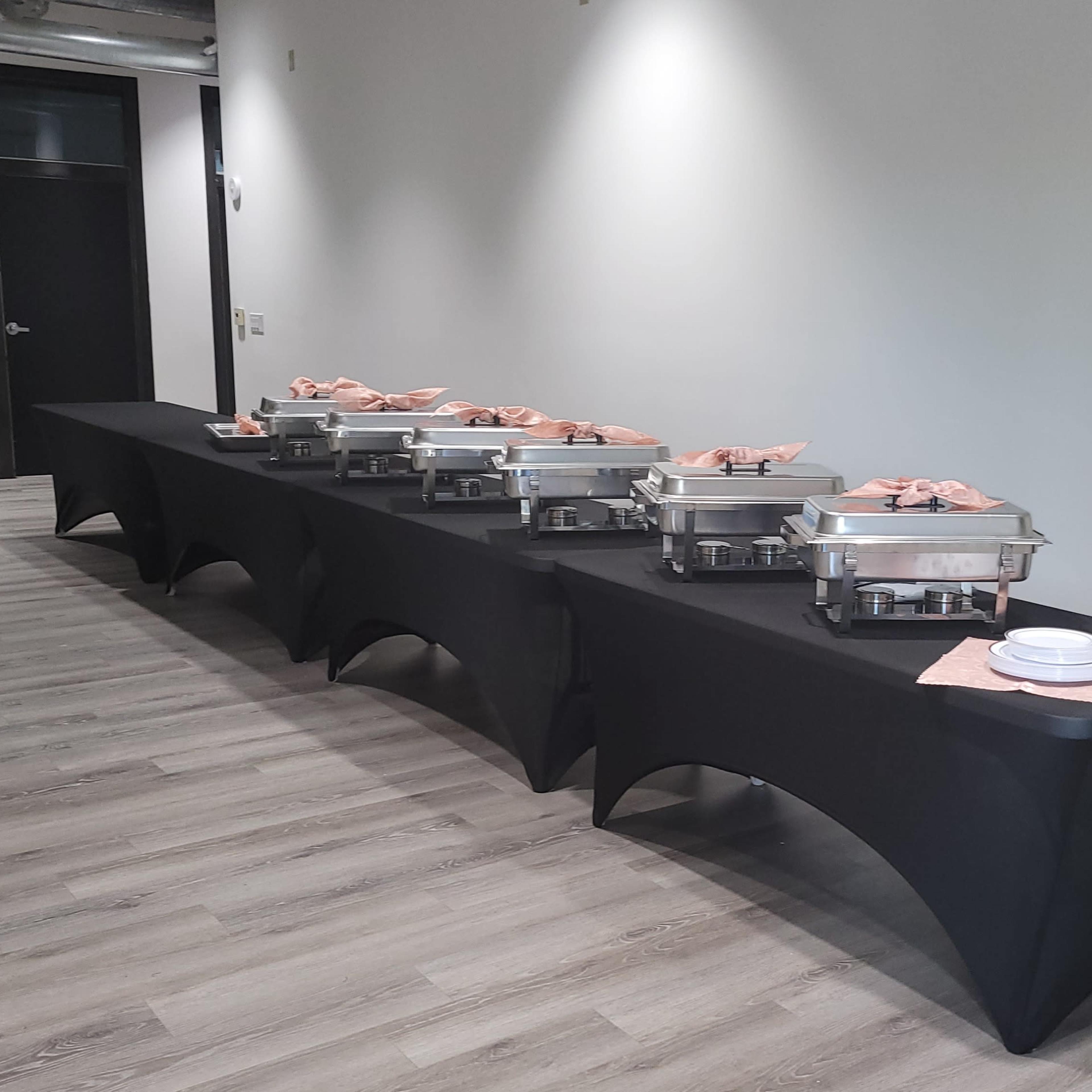 A row of chafing dishes with food covers is set up on black tablecloths in a modern hallway.