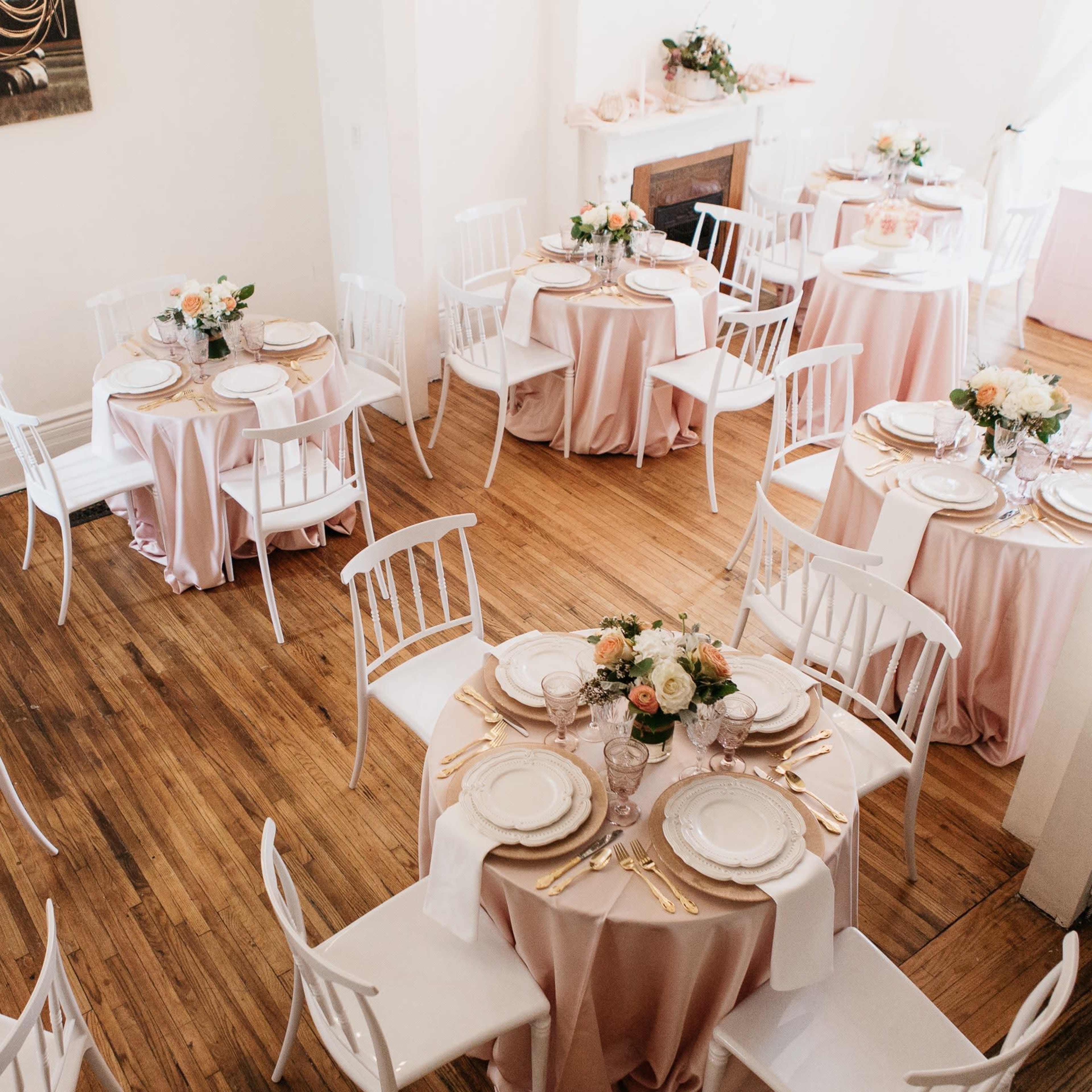 A brightly lit room features several elegantly set tables covered with pink tablecloths and adorned with floral centerpieces.