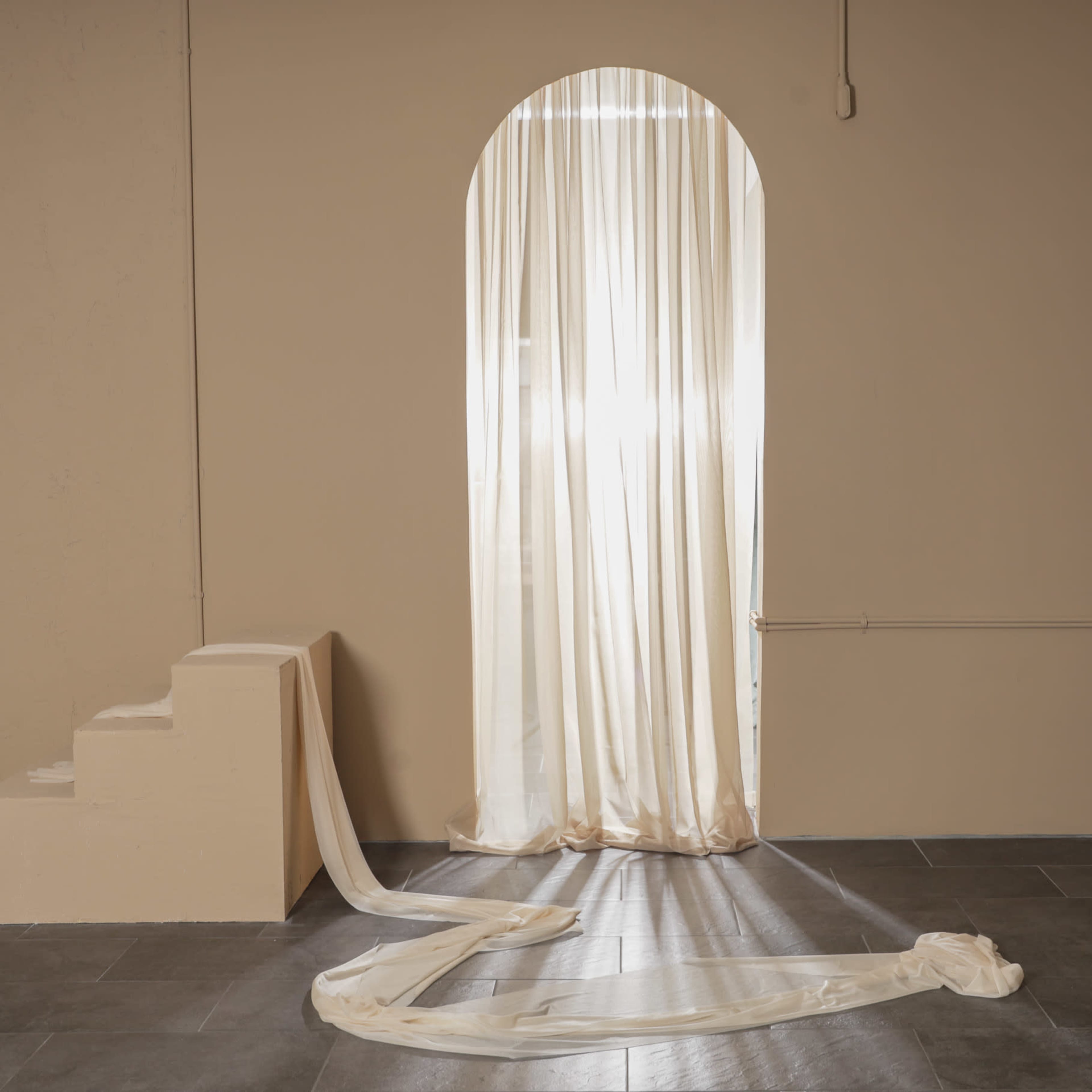 A sheer curtain drapes over an arched doorway, with soft fabric cascading along the floor beside a small staircase in a minimalist room.