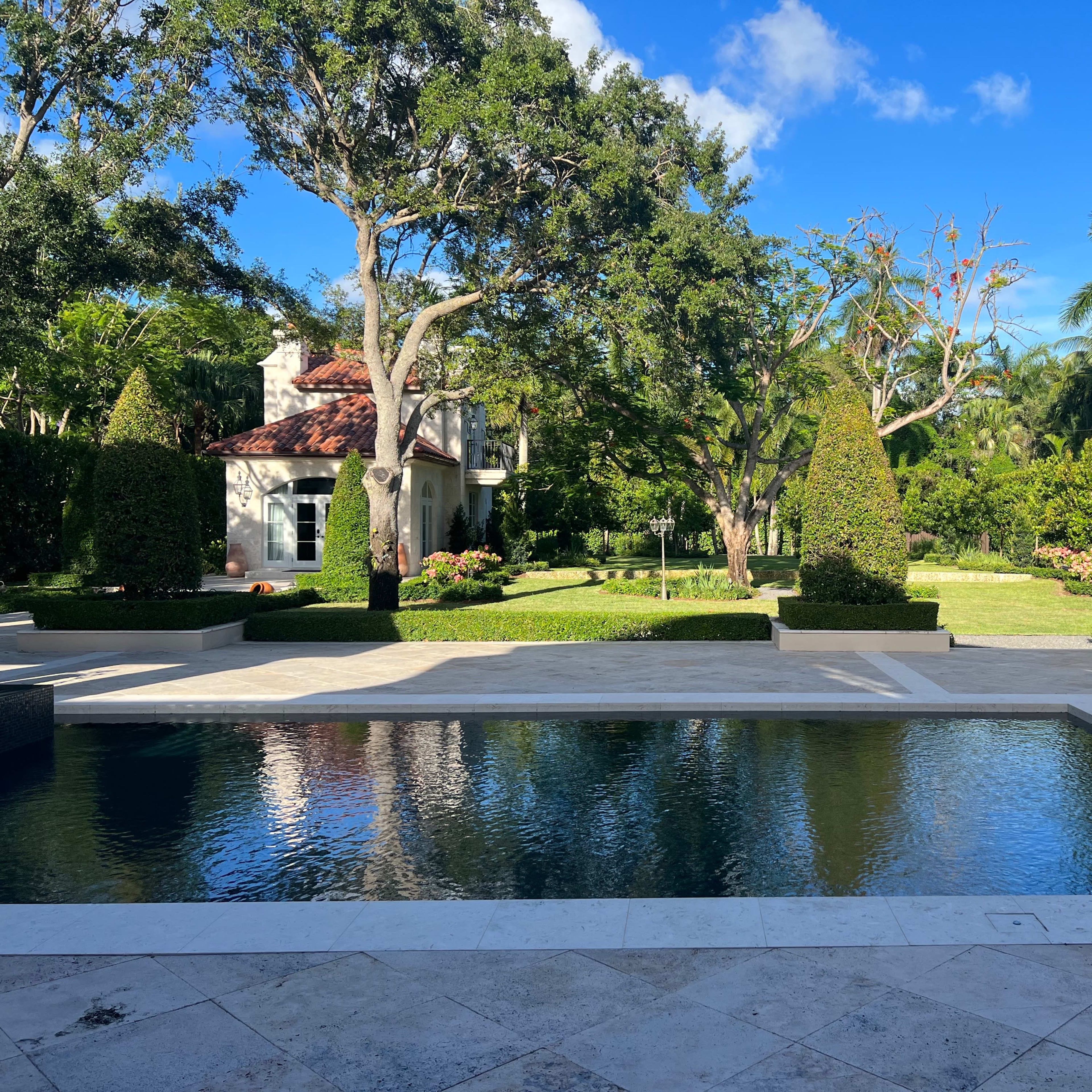 A landscaped backyard featuring a stone pool surrounded by lush greenery and trees, with a house visible on the right.