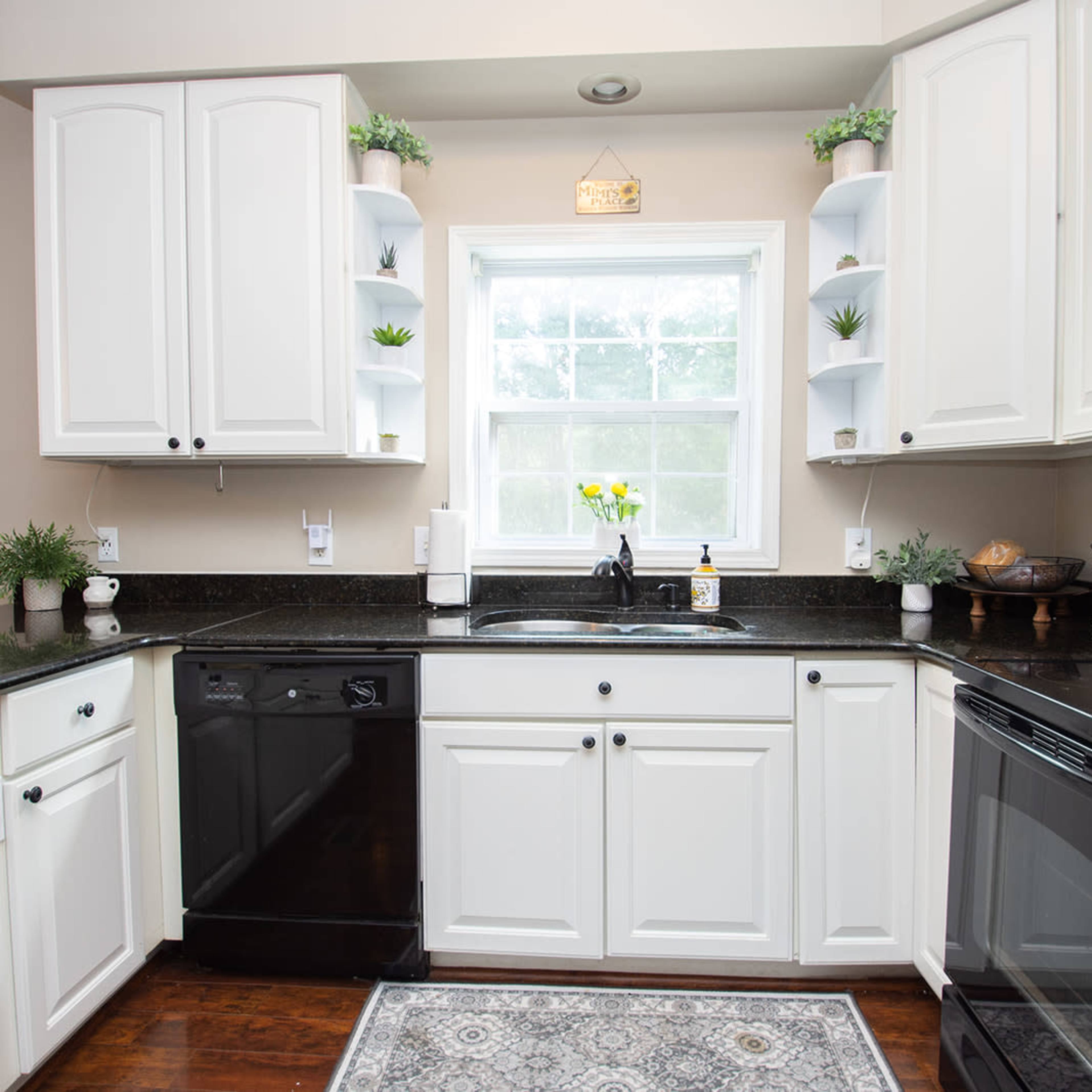 A modern kitchen with white cabinets, a dark countertop, and a window above a sink surrounded by plants and small decorative items.