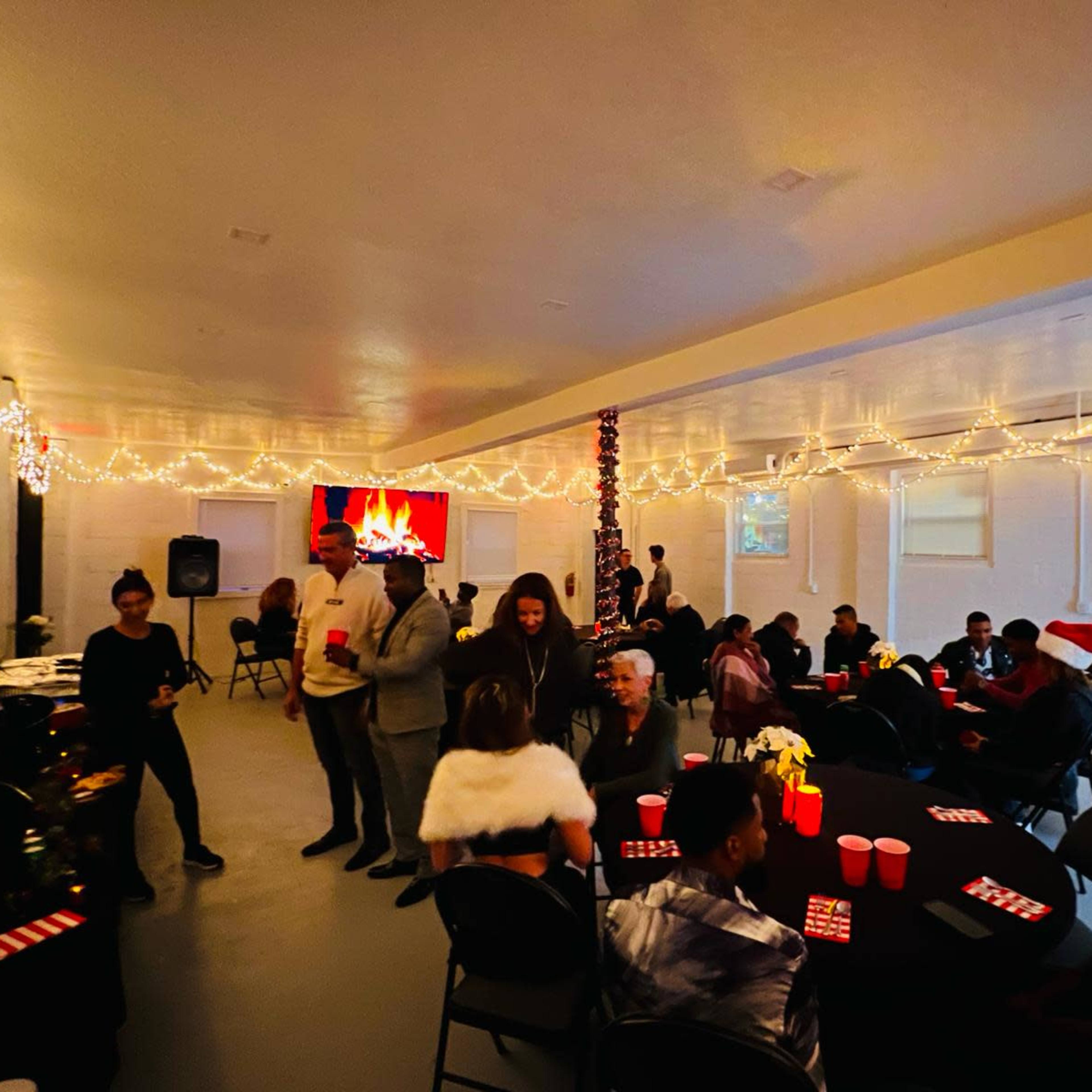 A decorated indoor venue filled with people socializing around tables, featuring festive lighting and a screen displaying a fireplace image.