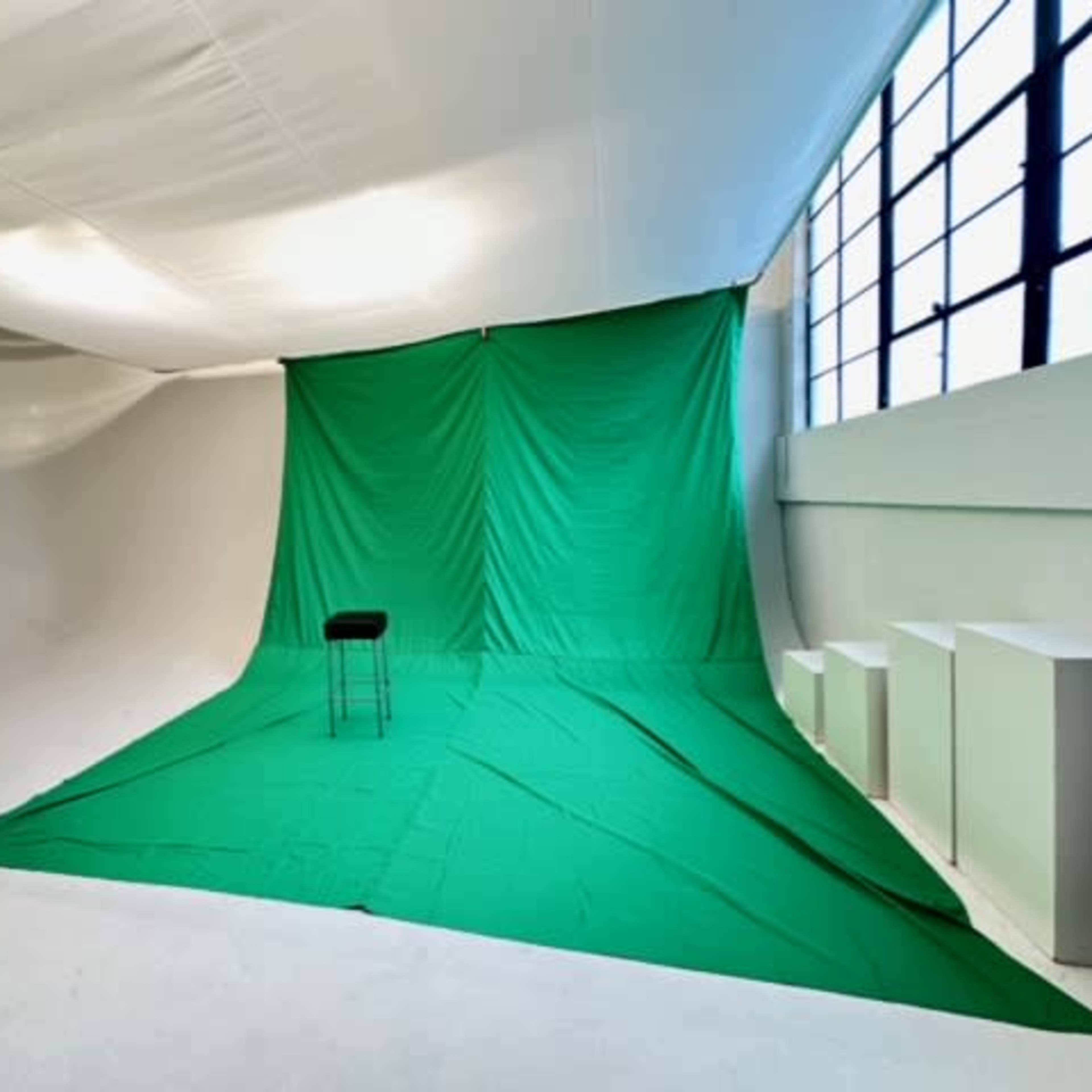 A green backdrop with a black stool is set up in a spacious studio with white walls and large windows.