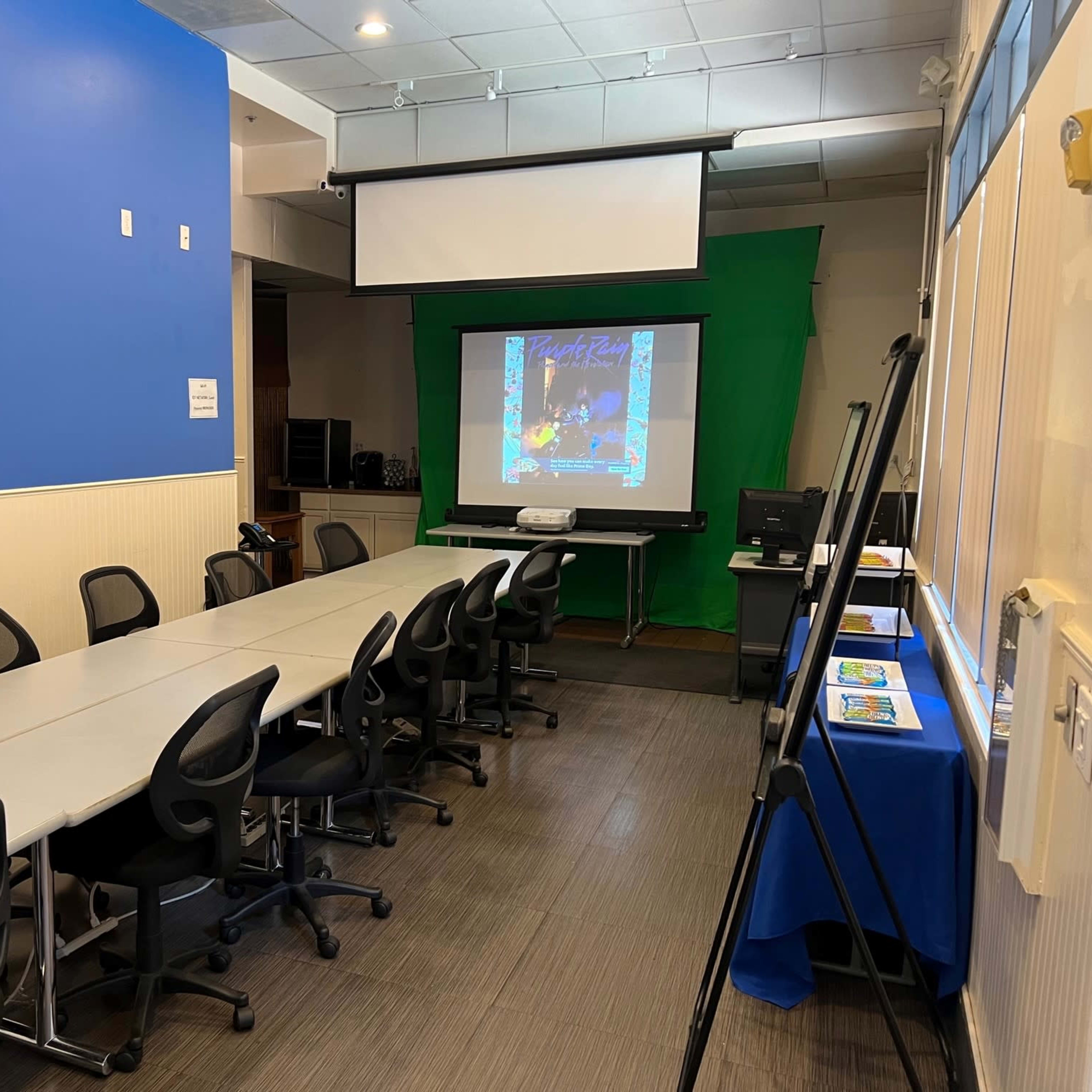 A conference room features a long table with chairs, a projector screen against a green backdrop, and a display table with promotional materials.