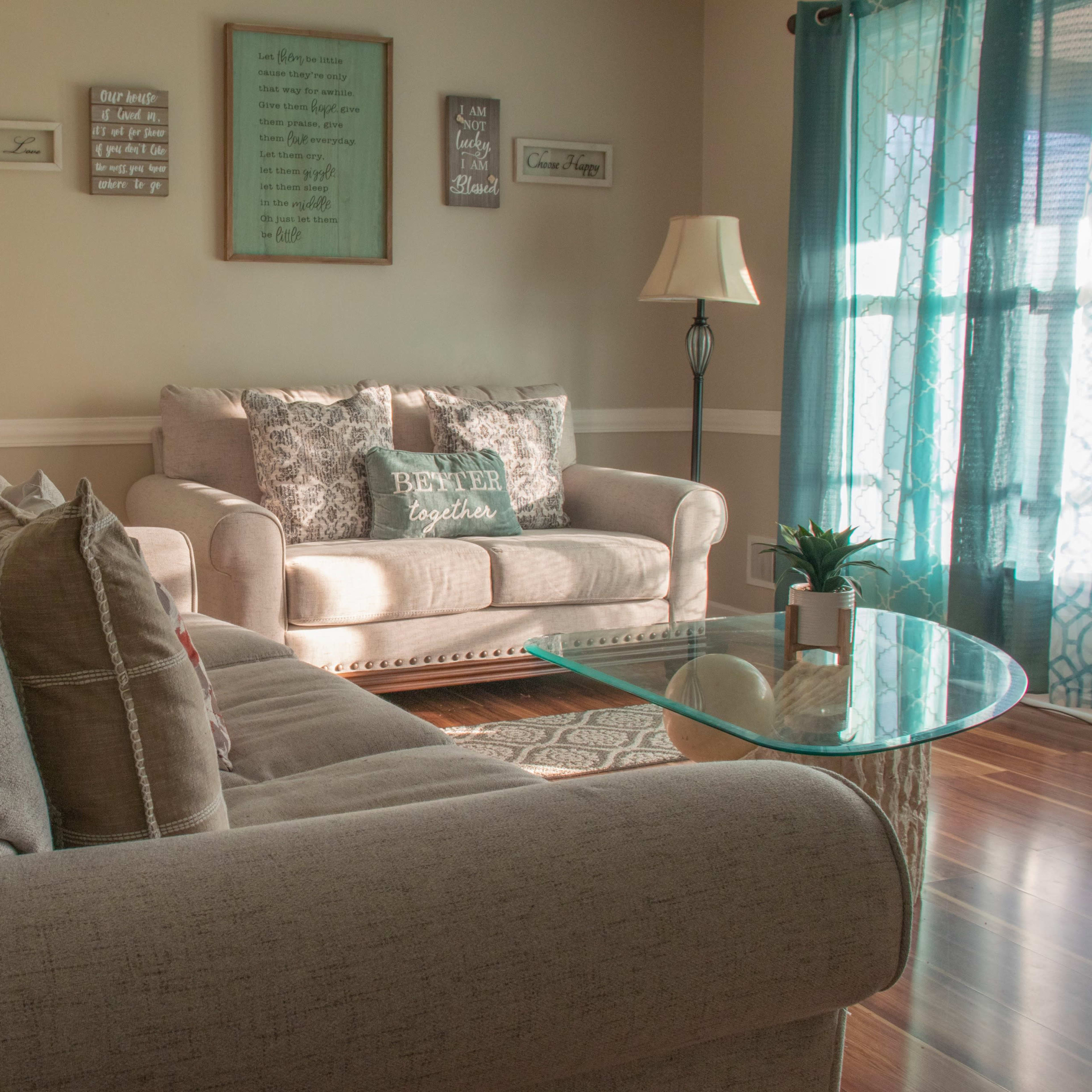 A living room features a light-colored sofa and armchair, a glass coffee table, and decorative wall art, with sunlight streaming through a window with sheer curtains.