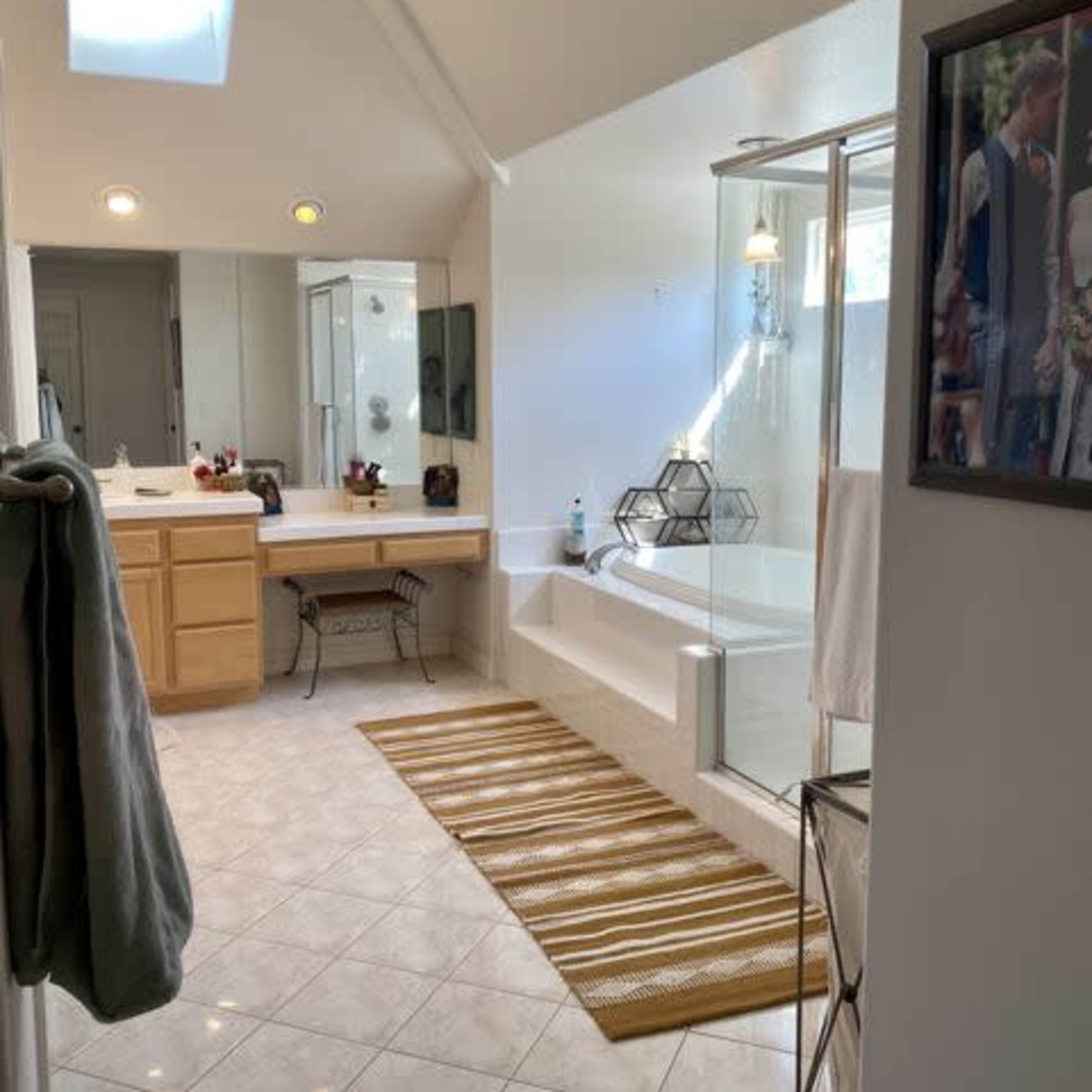 A bright bathroom featuring a large bathtub, a glass shower, a vanity with a mirror, and a decorative rug on the tiled floor.