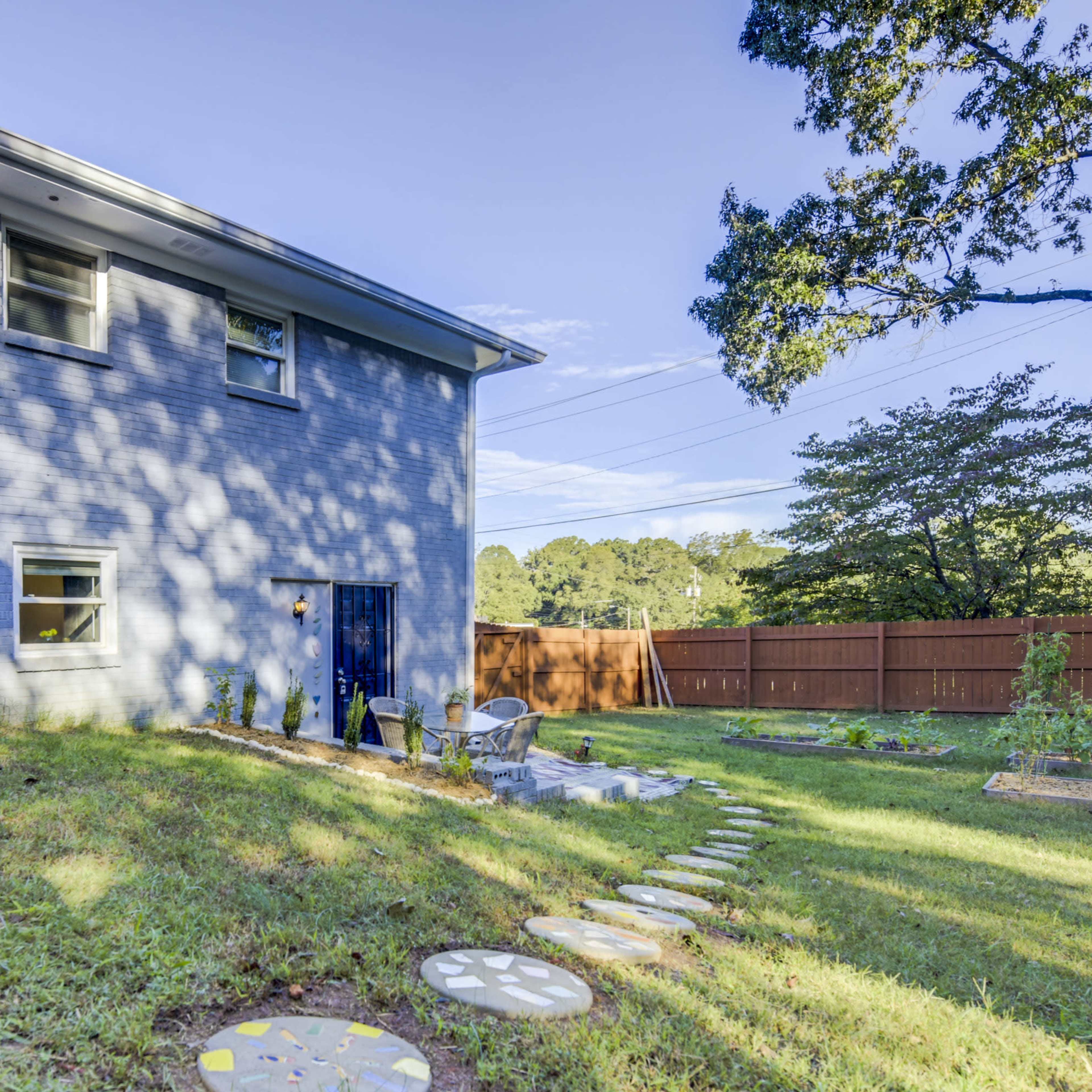 A two-story blue house is situated in a yard with a stone path leading to a patio area surrounded by plants and trees.