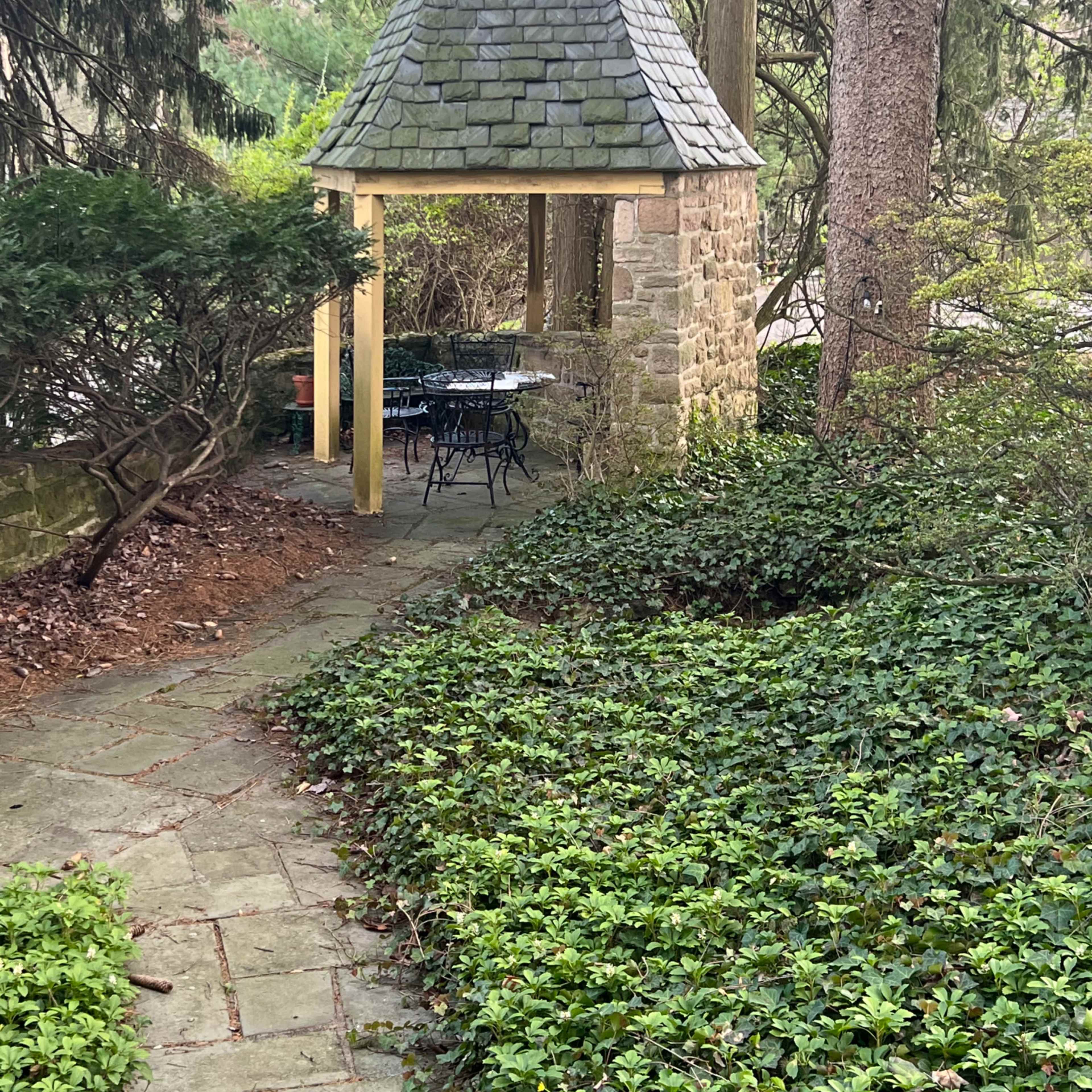 A stone gazebo with a conical roof is situated along a paved path, surrounded by lush greenery and trees.
