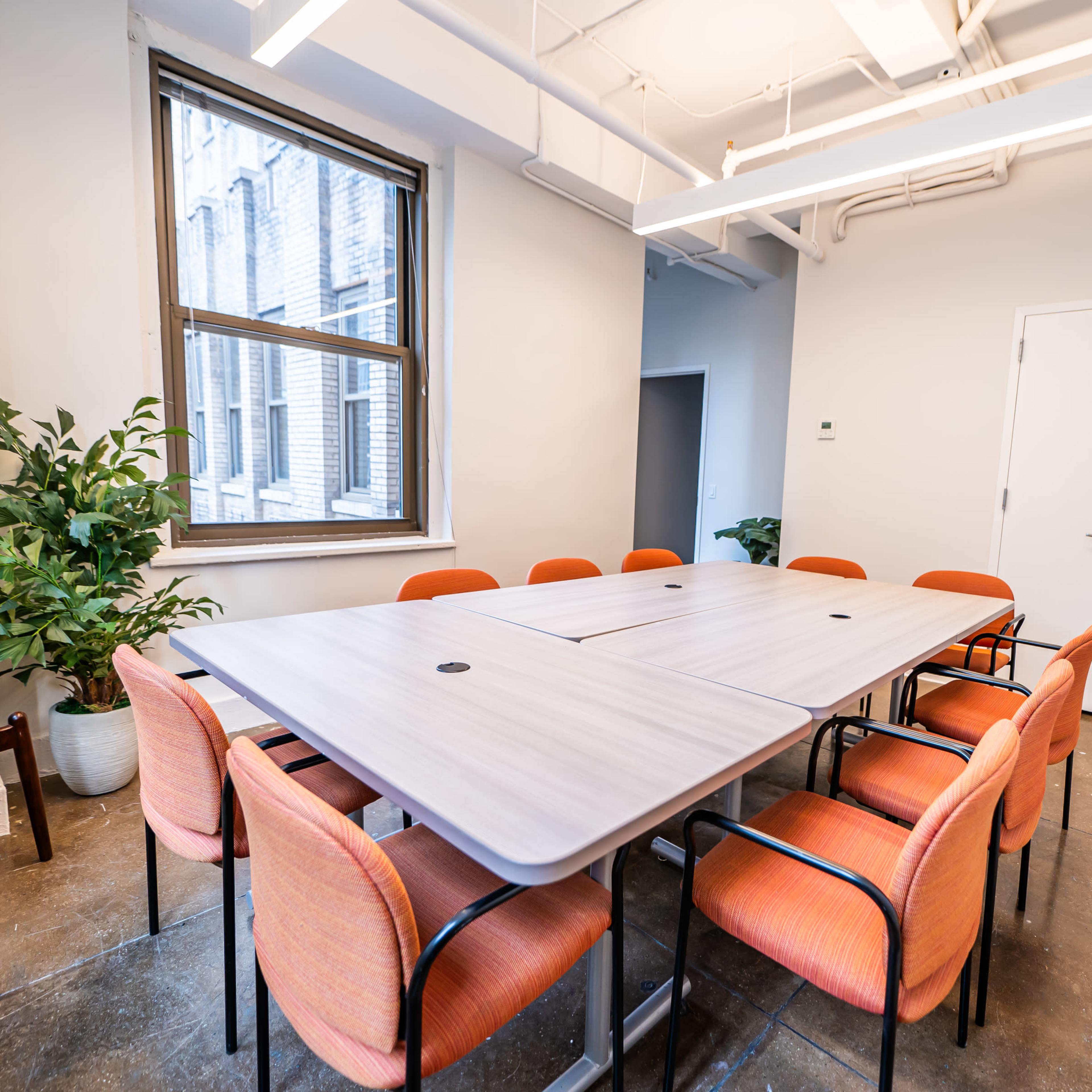 A conference room features a large rectangular table surrounded by orange chairs, with natural light coming through a window and potted plants in the corners.