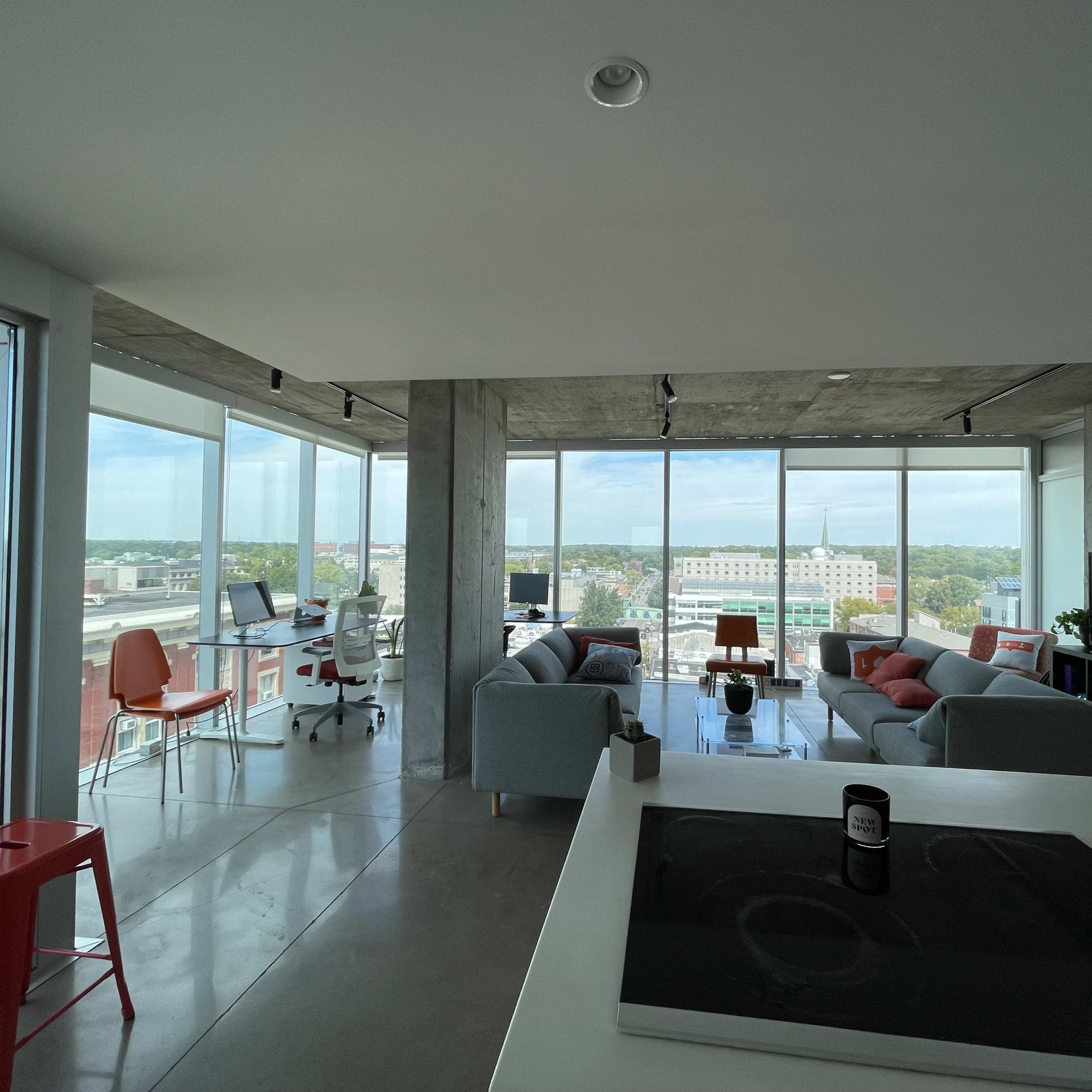A modern open-concept living space with large windows, offering a view of the city and featuring a kitchen area, comfortable seating, and workstations.