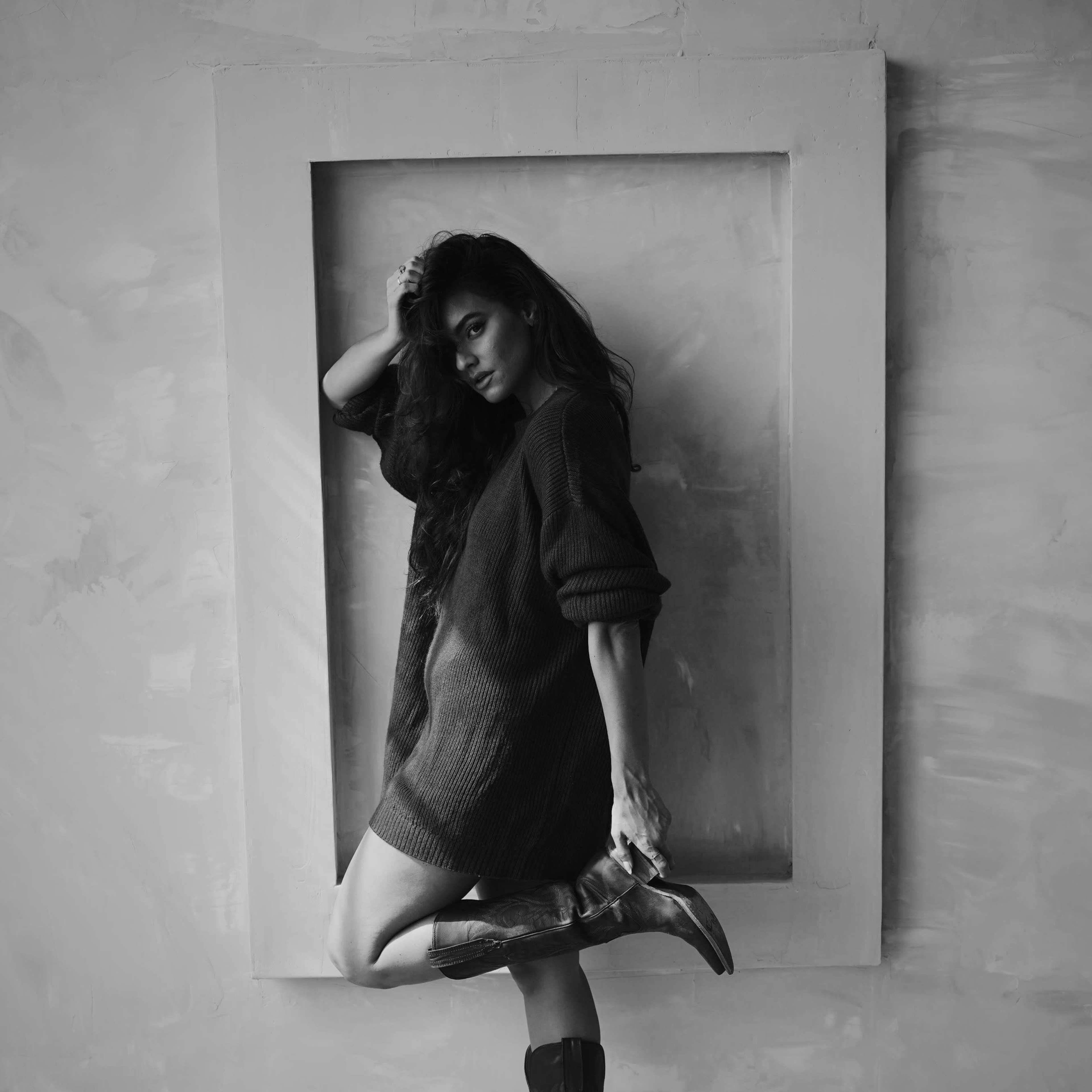 A woman poses in a dark sweater and cowboy boots against a textured wall framed by a white rectangular outline.